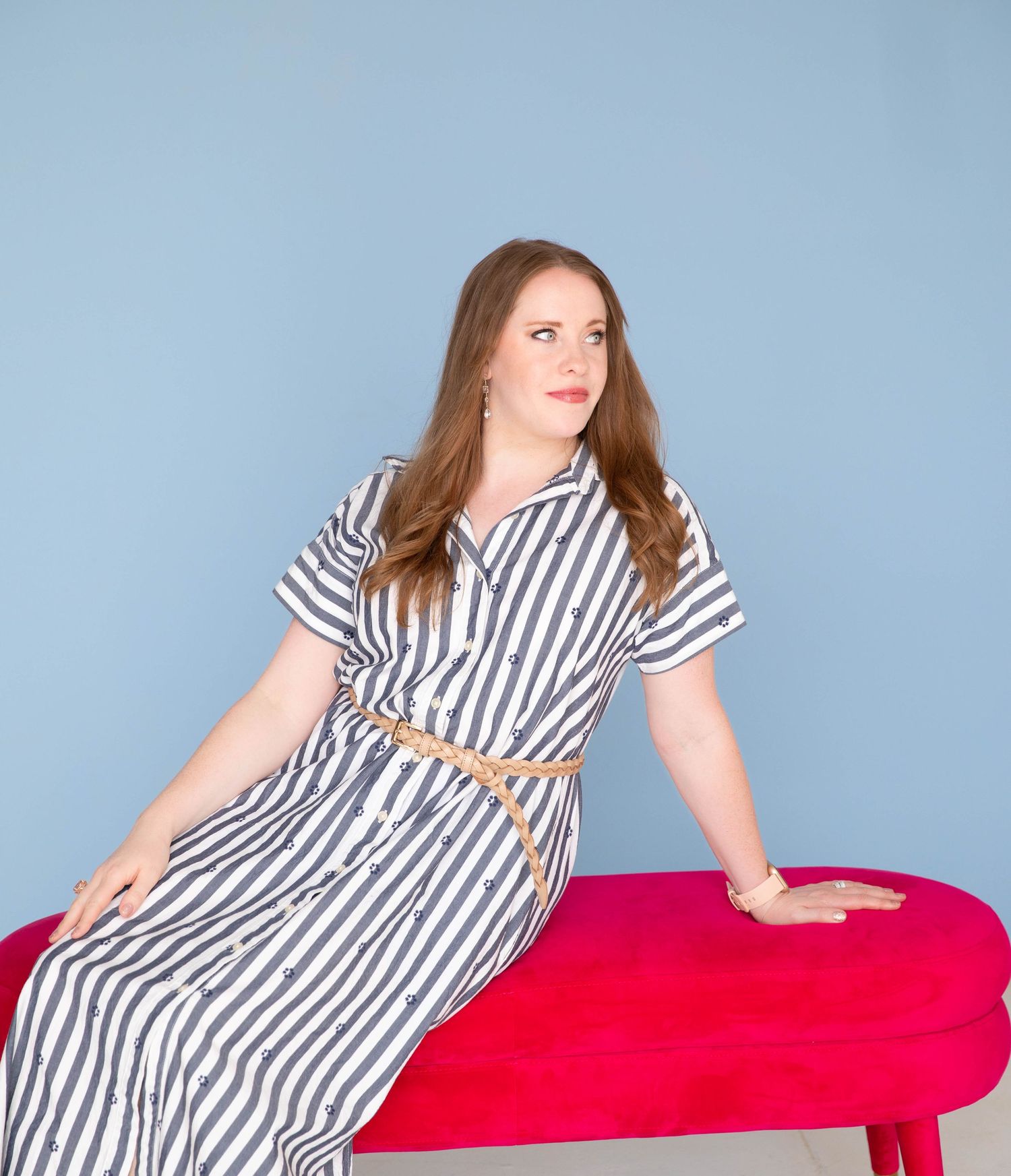 Person in black and white striped dress with brown belt poses on red ottoman against light blue background.