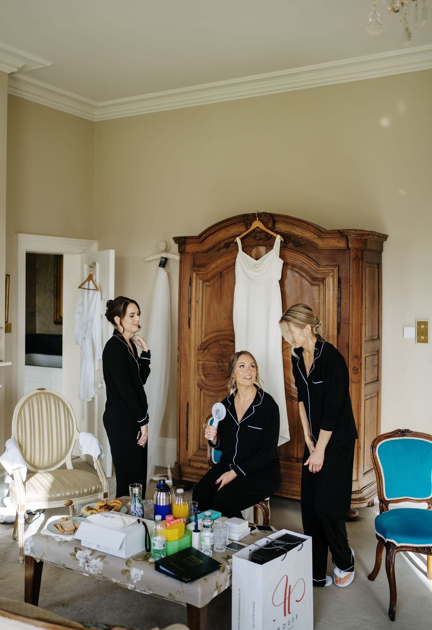 Bridal preparations in an elegant room with a white wedding dress hanging on an antique wooden armoire.