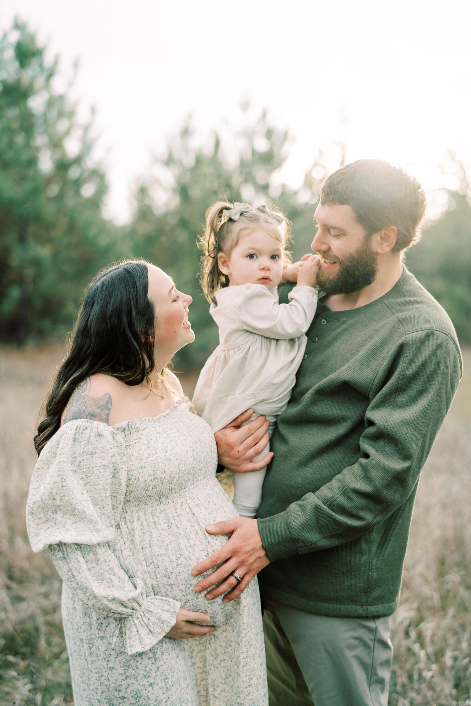 Family maternity photo in field with parents and toddler sharing tender moment at golden hour.