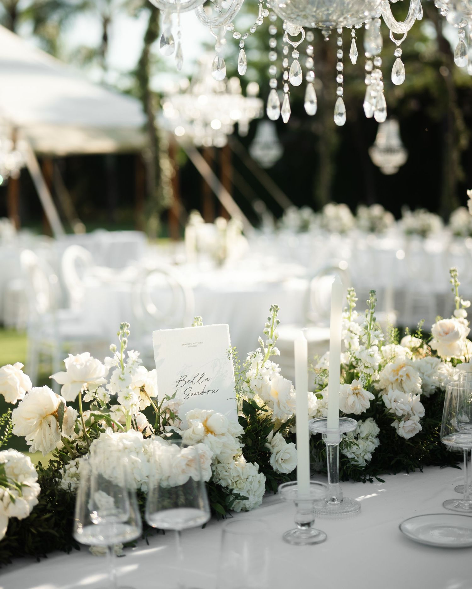 Elegant wedding table setting with crystal chandeliers, white floral arrangements and candles under an outdoor tent.