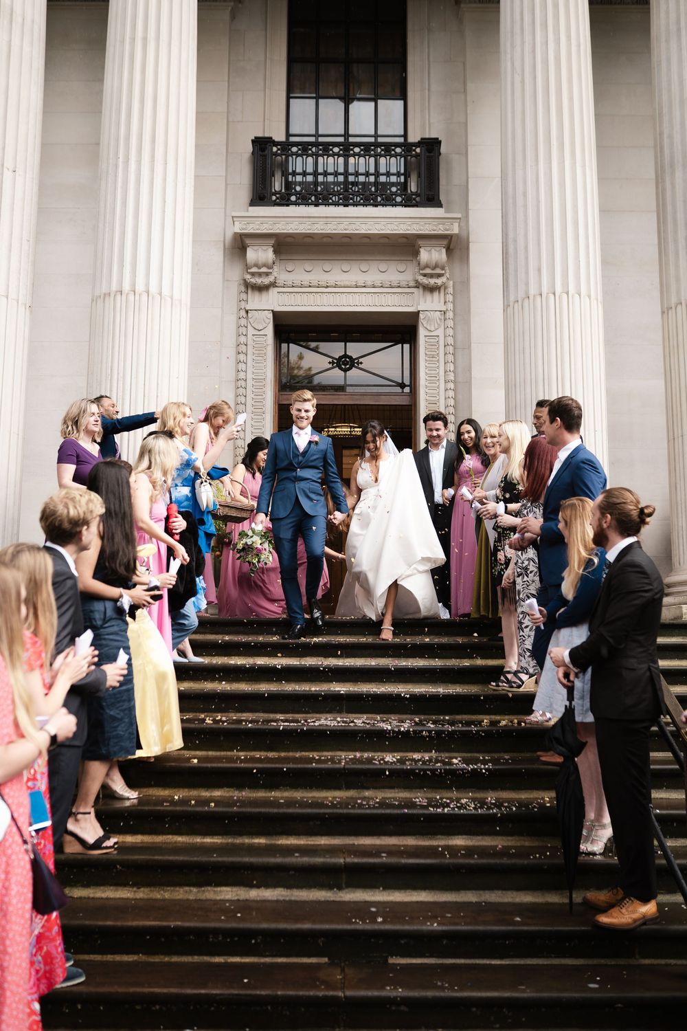 Wedding party gathered on grand stone steps with white columns for post-ceremony celebration.