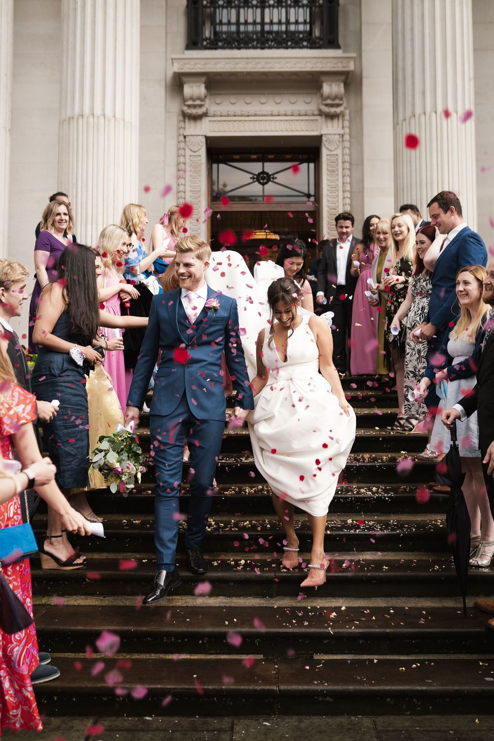 Newlyweds descending stone steps while guests throw rose petals in celebration.