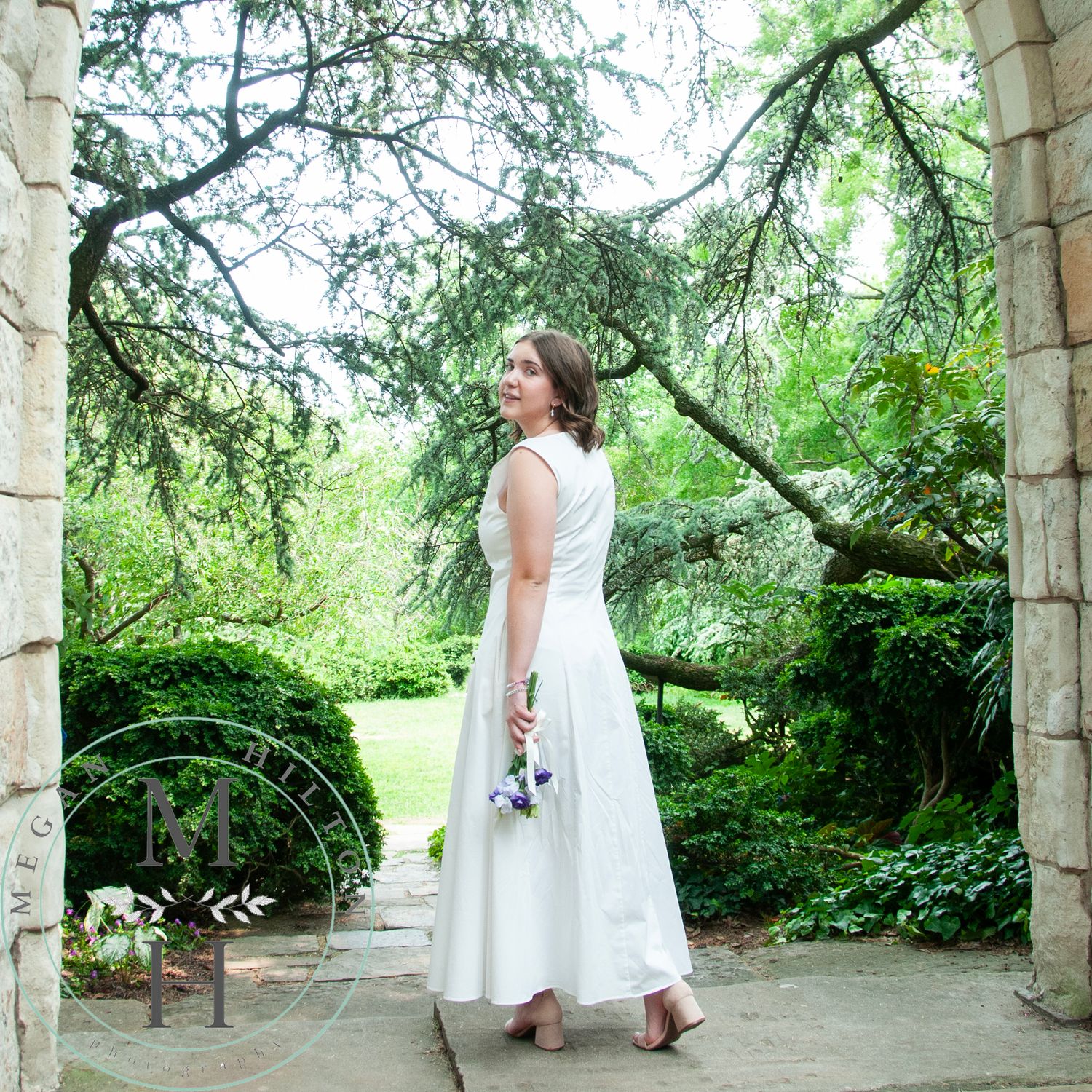 A full body photo of a young girl with a panoramic view of a garden pathway with stone columns and overhanging tree branches.