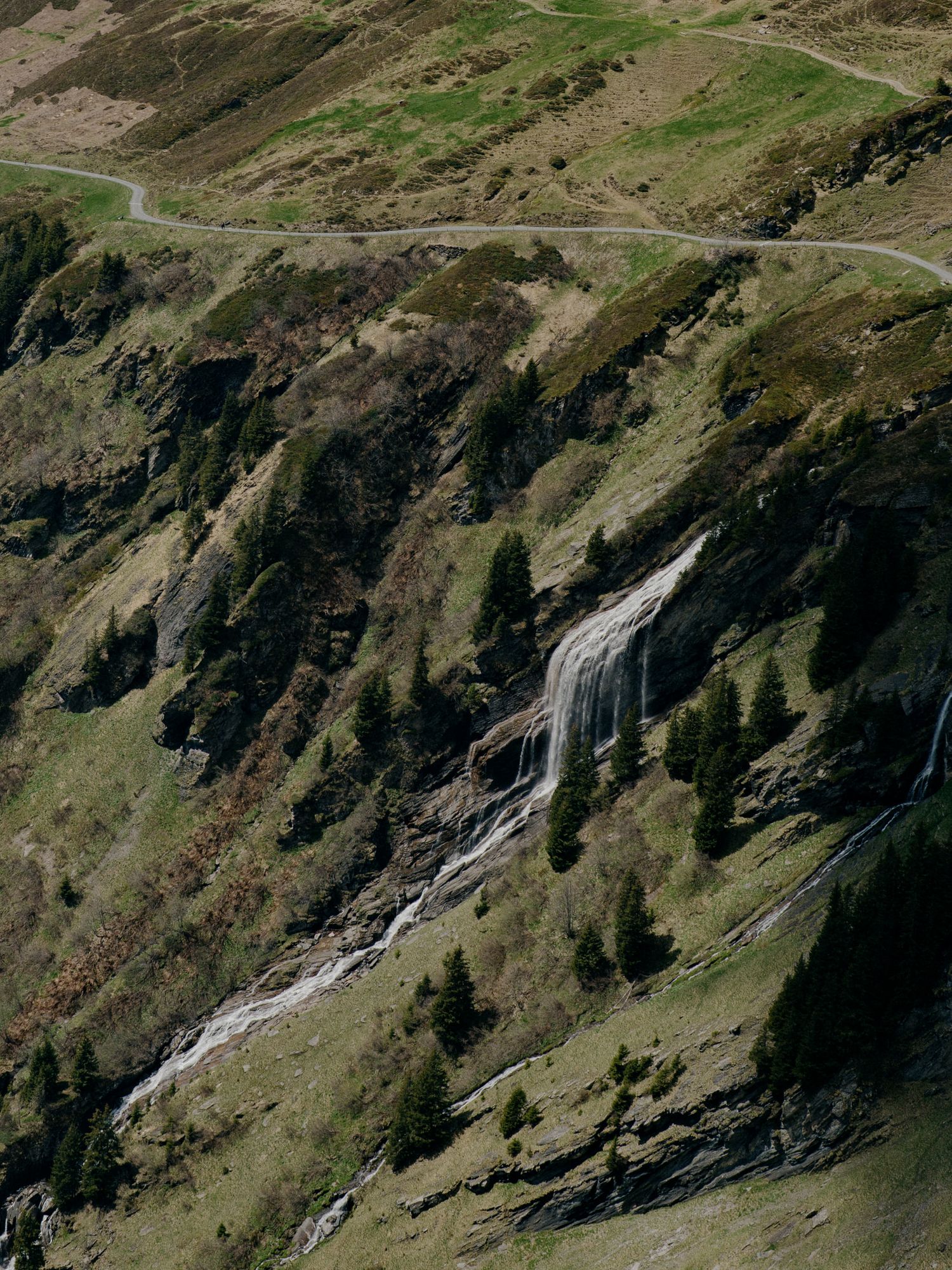 A winding mountain road winds past a cascading waterfall on a steep, rugged mountainside with scattered evergreen trees.