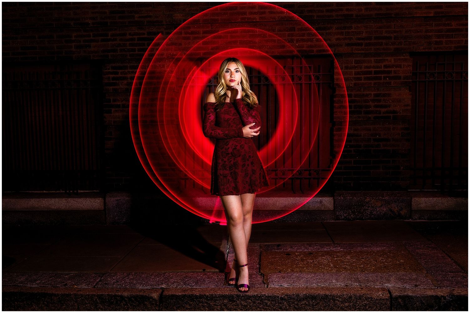A person in a burgundy dress poses against a brick wall with dramatic red circular lighting behind them.