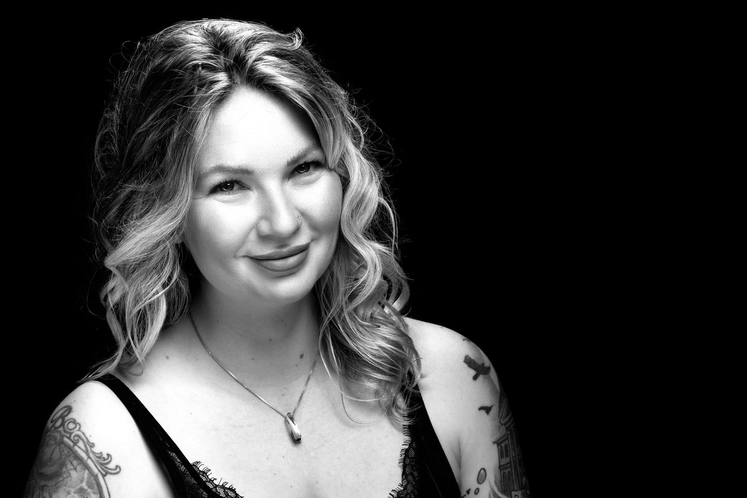 Black and white portrait shot with wavy blonde hair, wearing dark top and necklace against dark background.