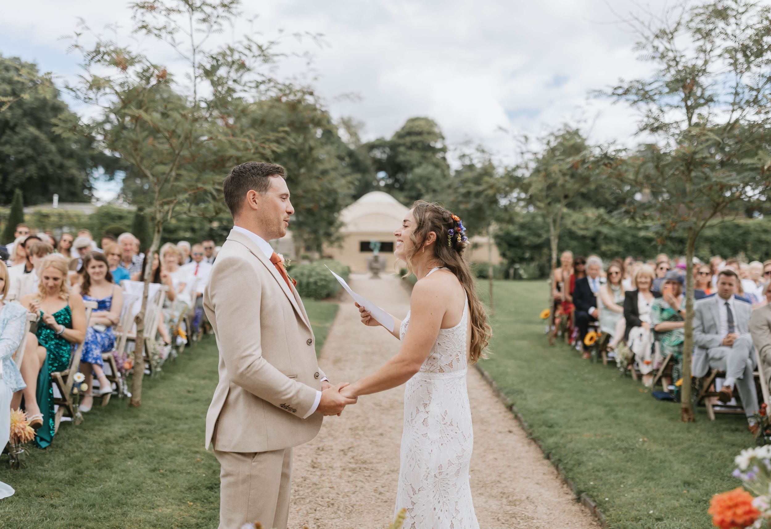 Couple exchanges vows during outdoor garden wedding ceremony with guests seated on lawn beneath cloudy sky.