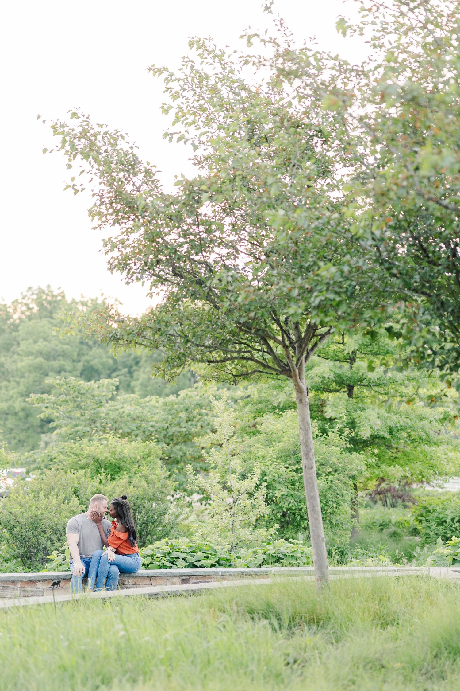 A couple sits together on a bench under a blossoming tree in a serene park setting on a bright summer day.