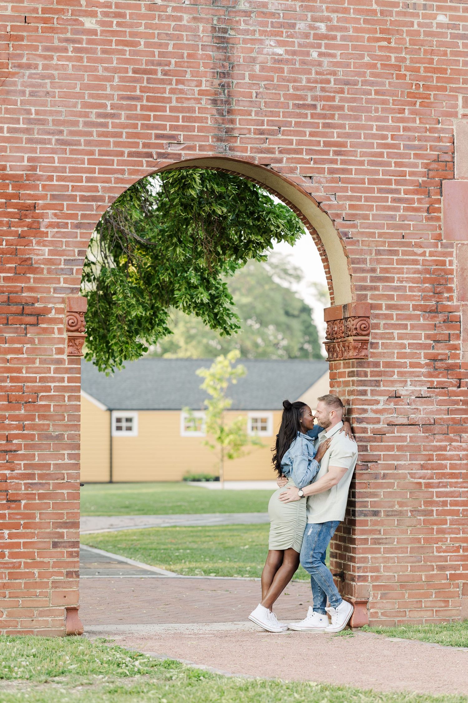 Couple sharing a romantic moment under a brick archway on a sunny day with green trees and buildings in the background.