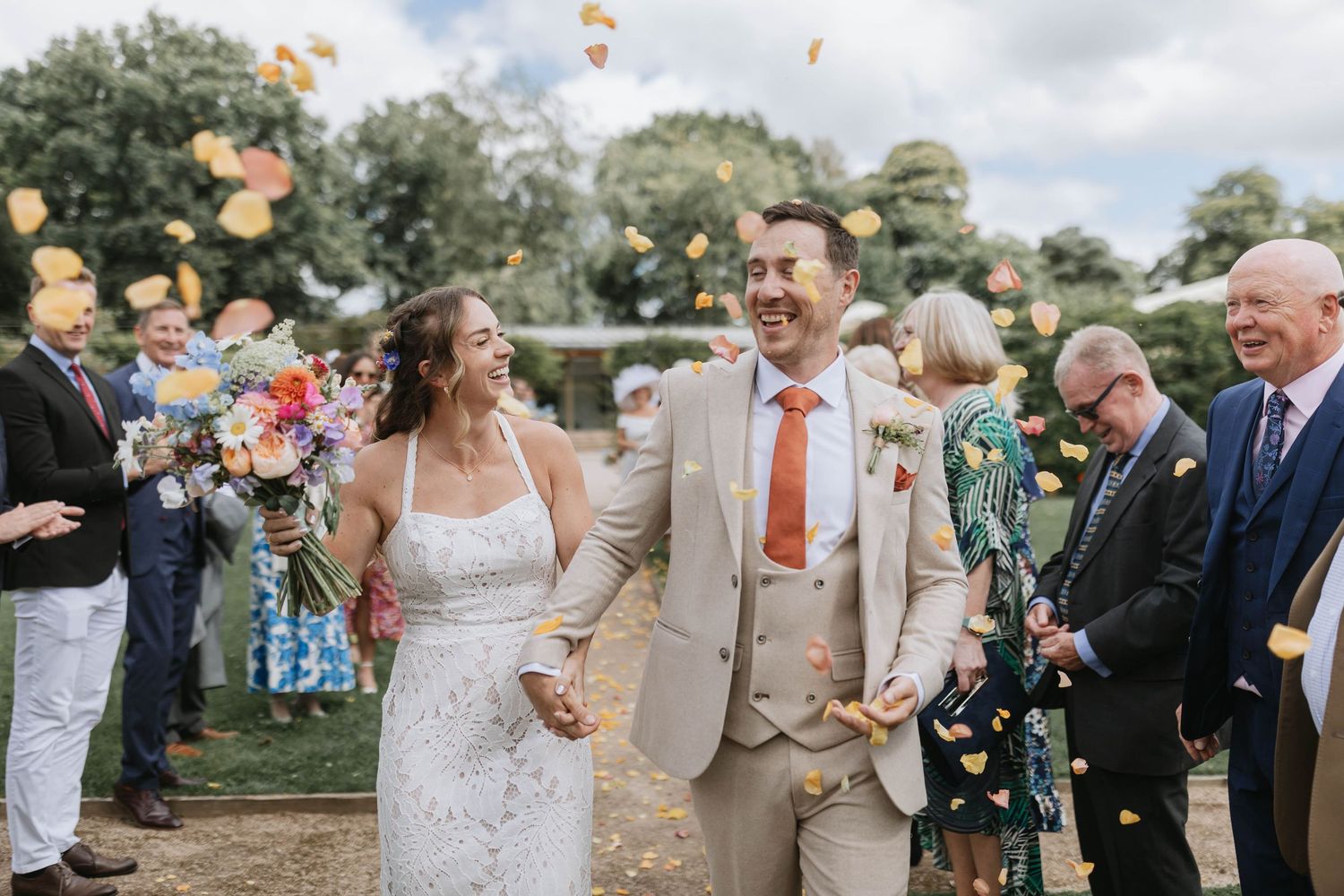 Wedding guests toss flower petals as newlyweds walk down grassy aisle at outdoor summer celebration.