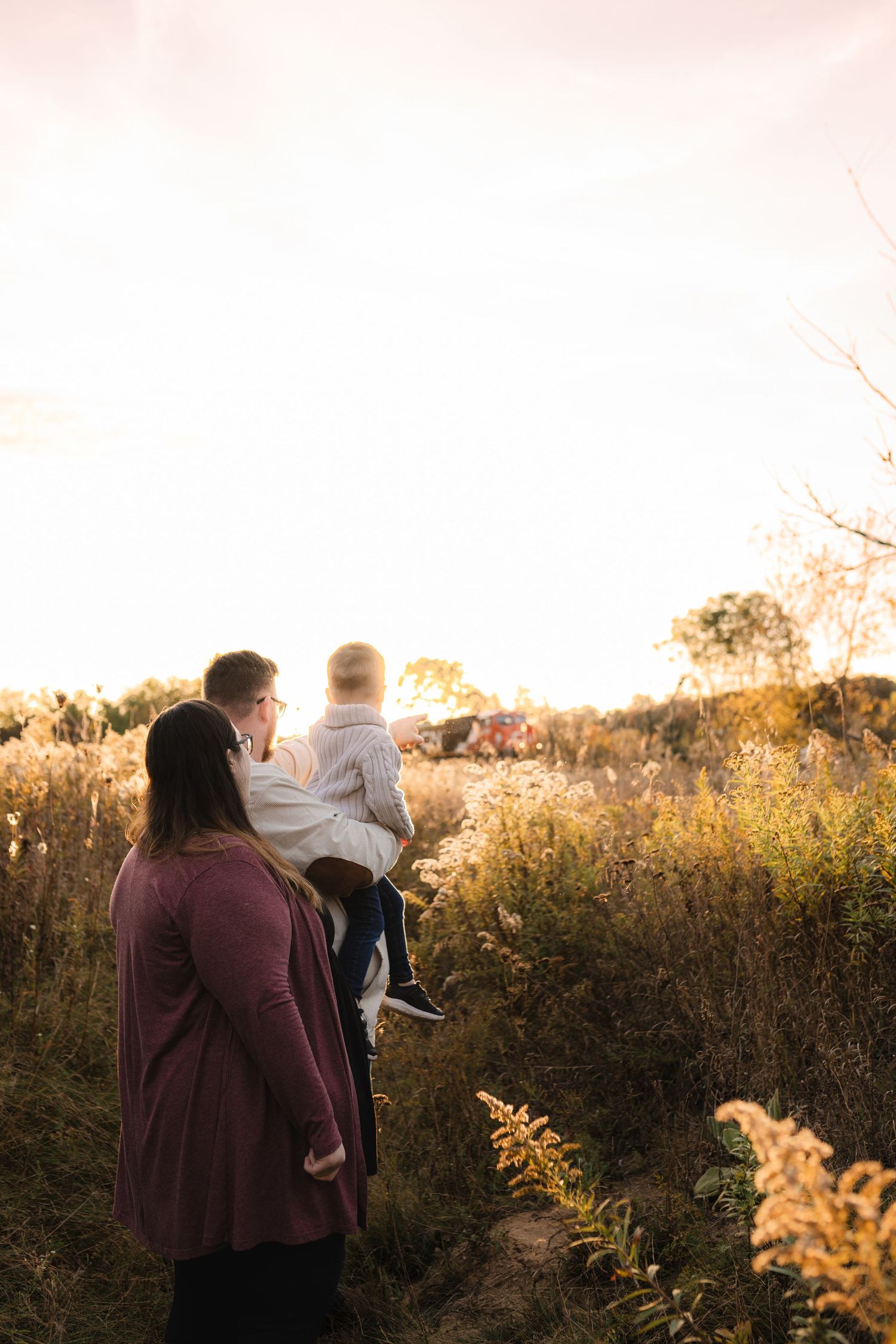 Silhouetted figures stand together in golden backlit field of tall grass during sunset, watching a train pass.