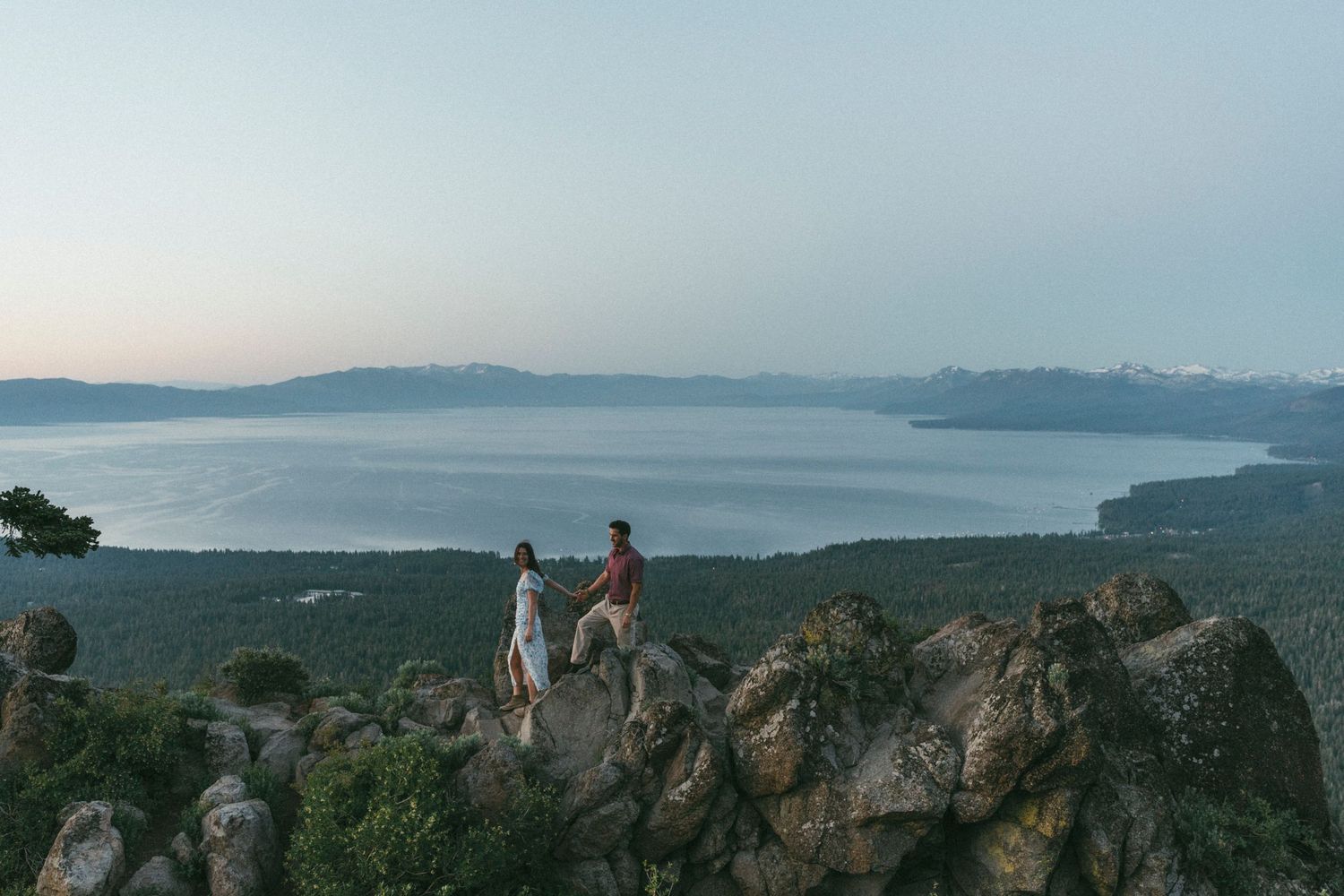A couple holds hands while standing on a rocky mountaintop overlooking a serene lake tahoe and mountain vista at dusk.