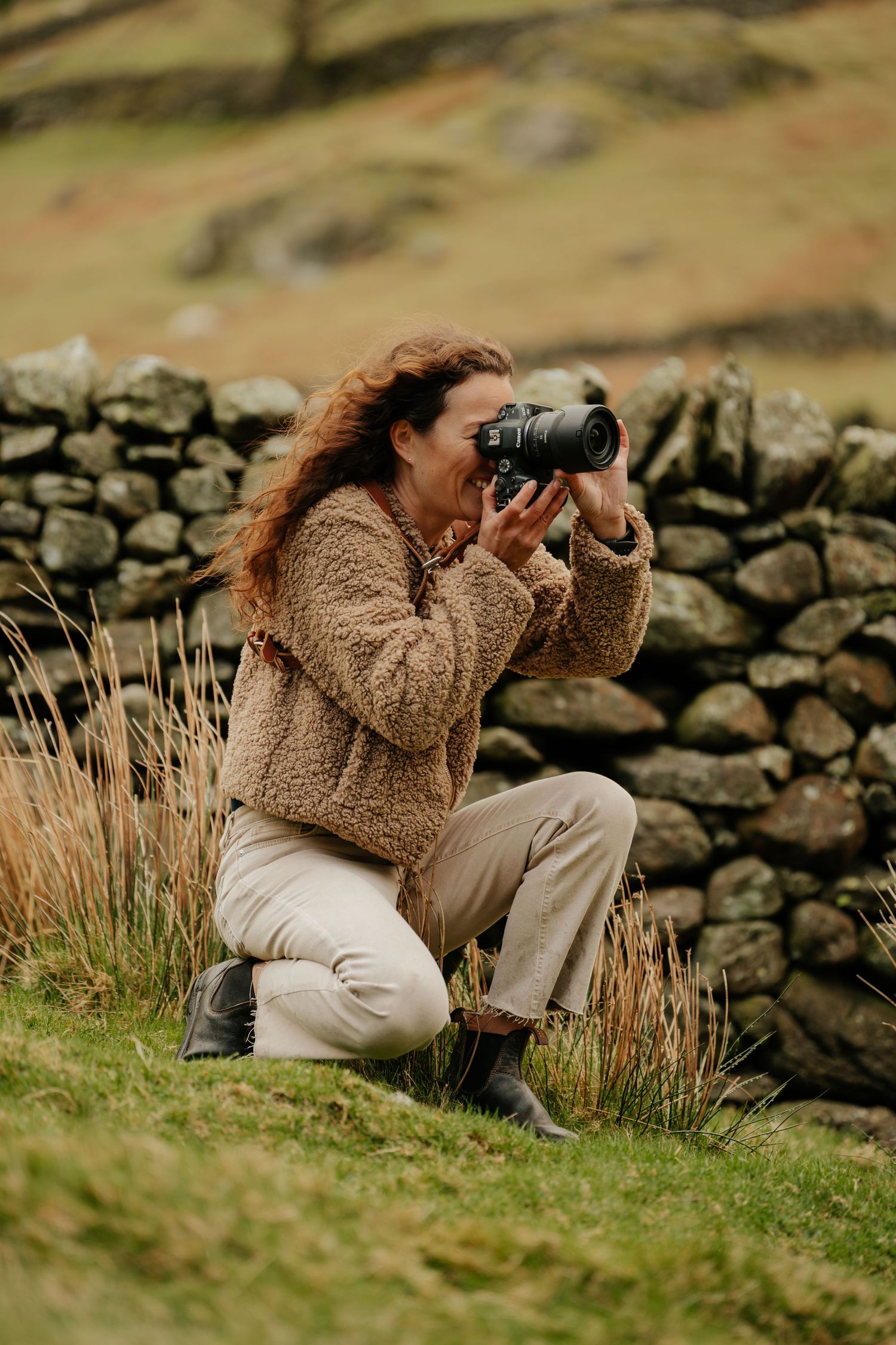 A photographer in a cozy sweater takes pictures while sitting by a stone wall in rolling hills.