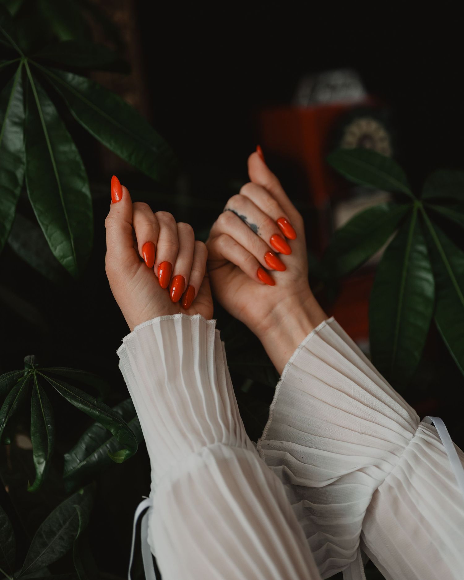 Hands with bright orange nail polish shown against dark green leaves and white sleeves in moody lighting.