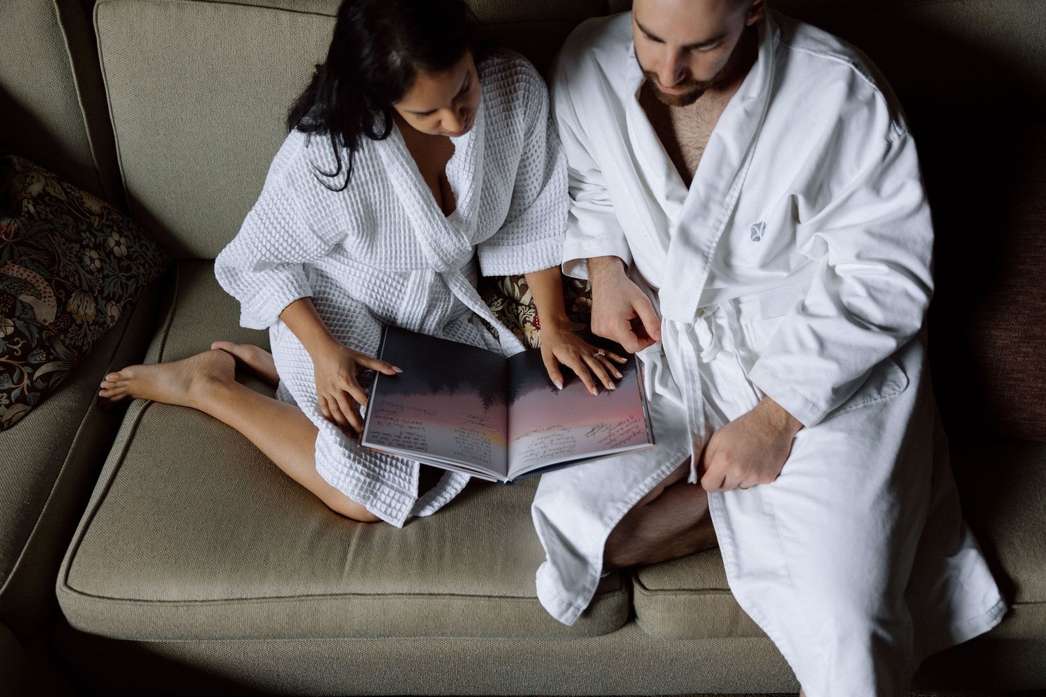 Two people in white bathrobes relax together on a beige couch while looking at documents.