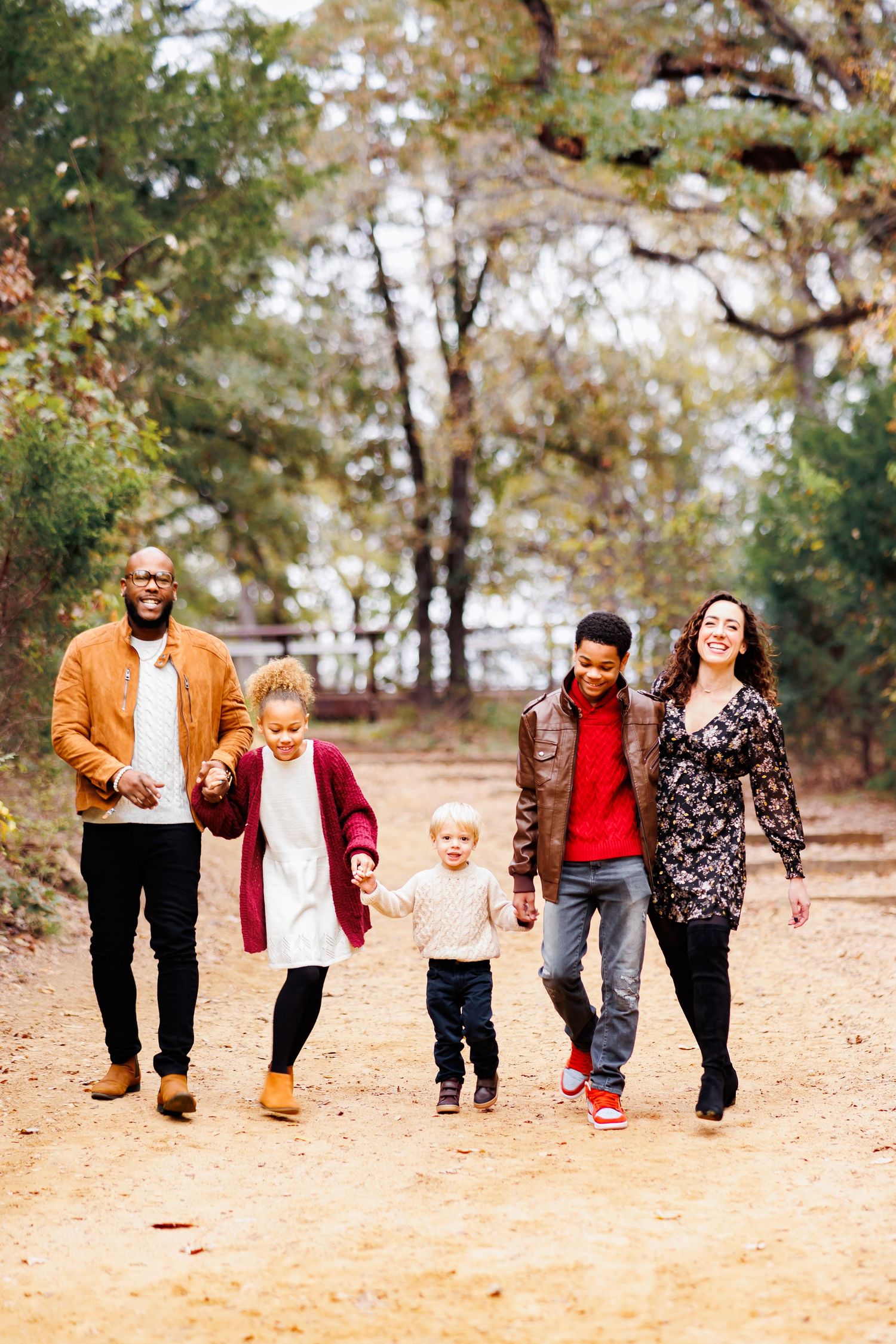A family holding hands and walking together through a sunlit autumn forest path in Dallas wearing coordinating burgundy and caramel outfits