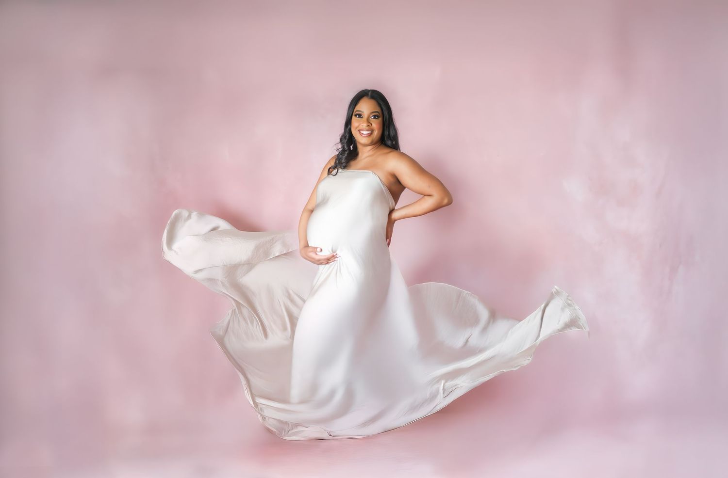 Artistic maternity photo showing a woman in flowing white gown against a pink backdrop in a Dallas studio