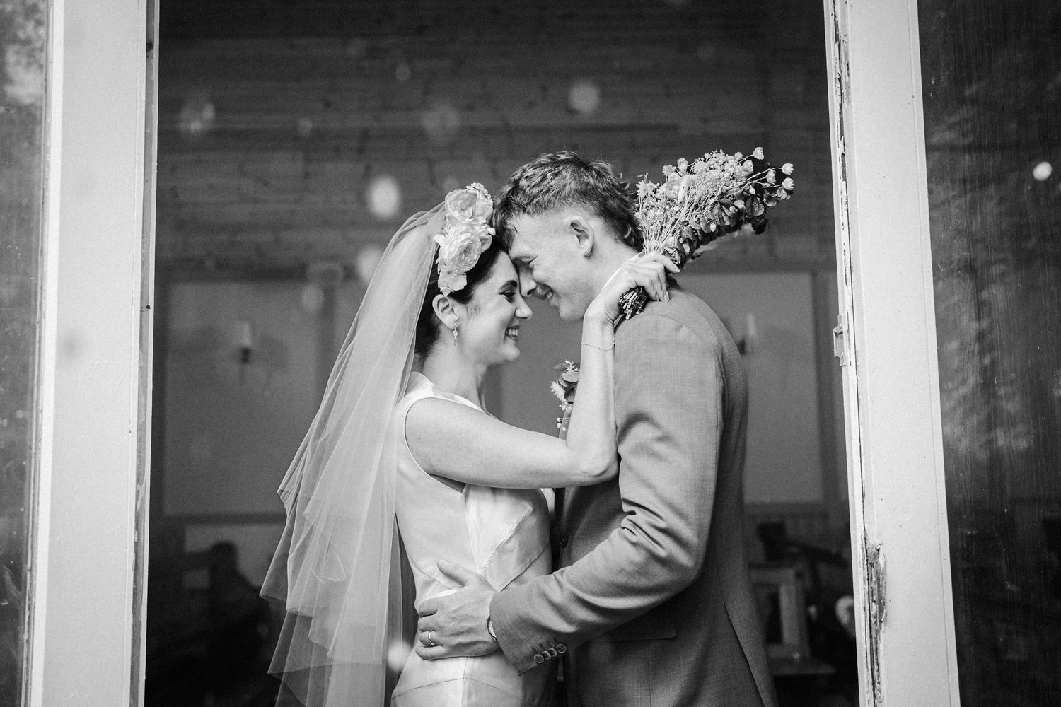 A tender moment between newlyweds sharing a romantic embrace in a rustic doorway captured in black and white photography.