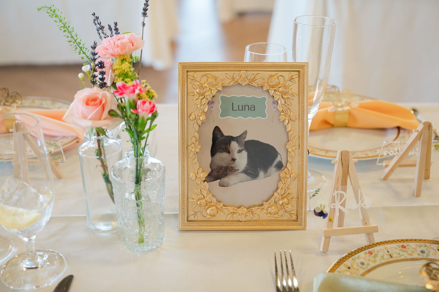 A collection of table decor at a wedding reception featuring floral centerpieces, gold frames, and wine glasses.
