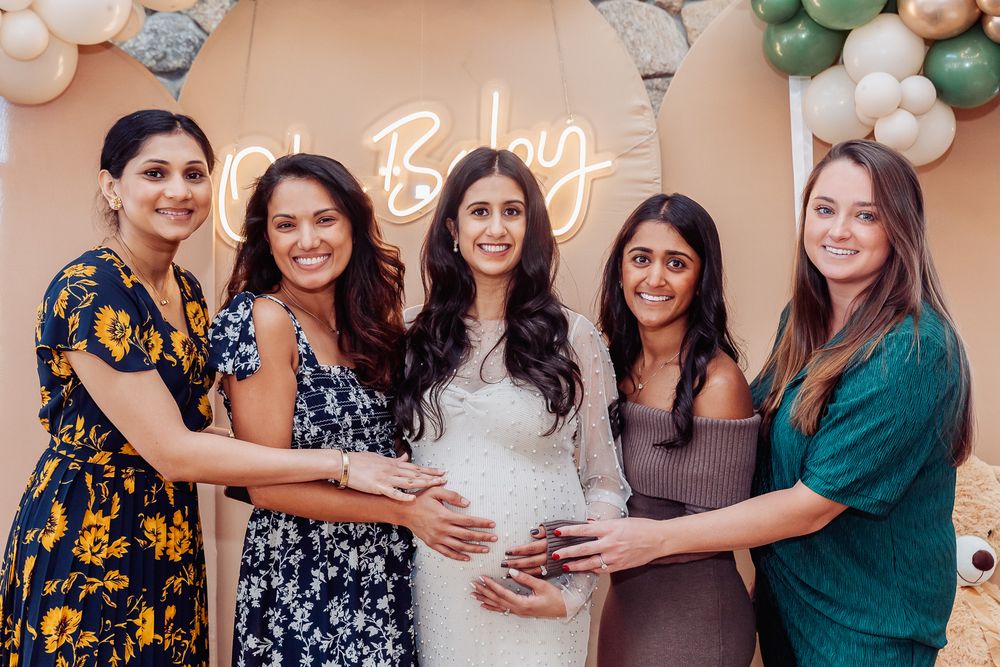 Women gather around for a baby shower photo in front of a decorative balloon arch with neon sign.
