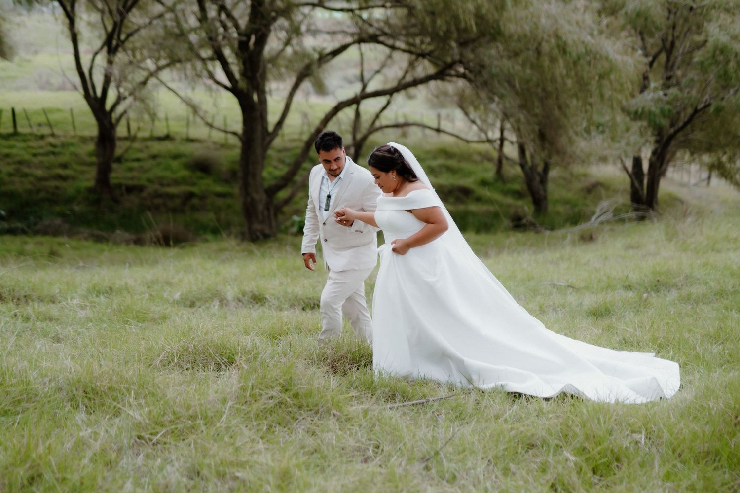 Couple walking through lush green meadow with flowing white wedding dress trailing behind.