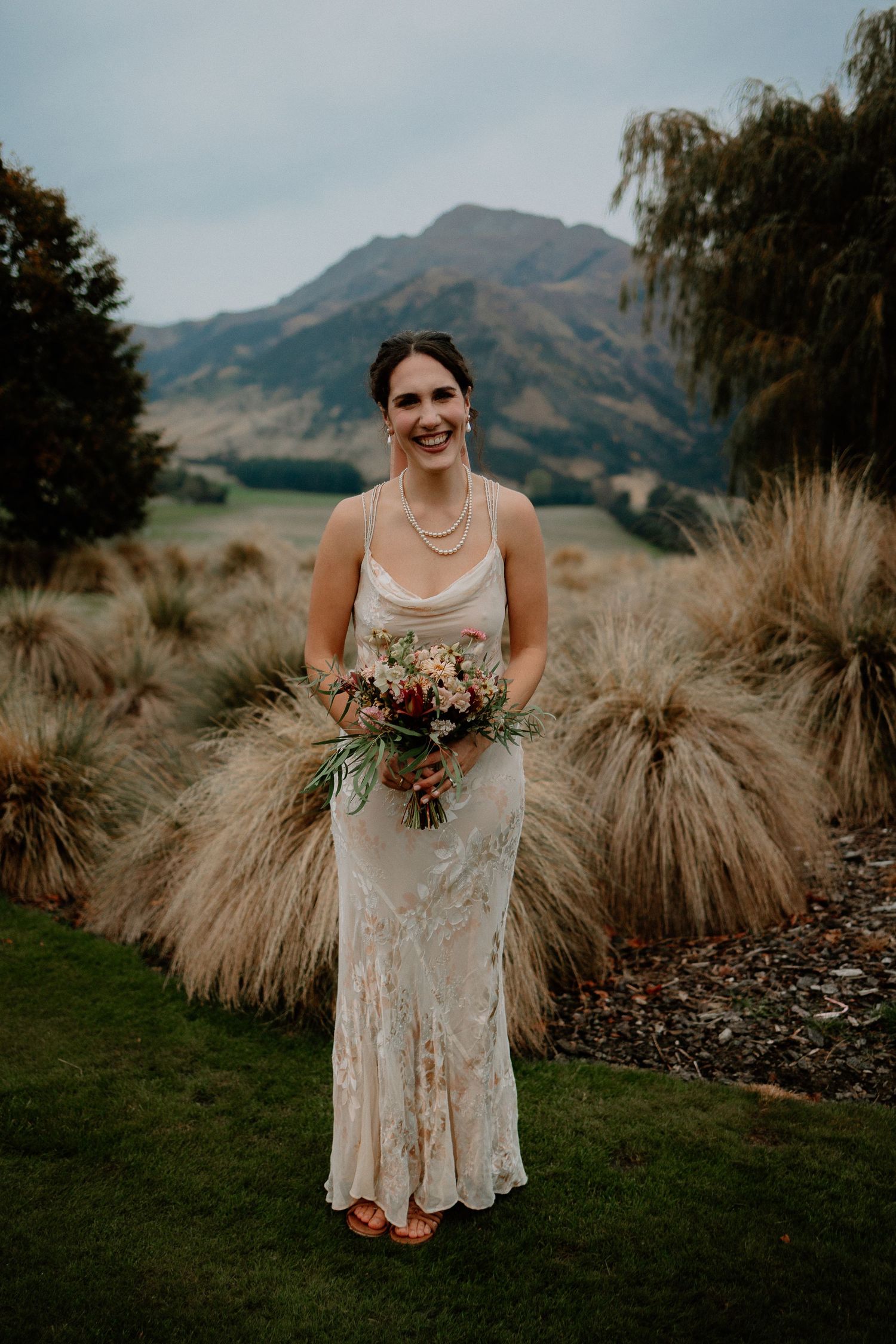 A bride in a fitted lace wedding dress stands among ornamental grasses with mountains in the background.