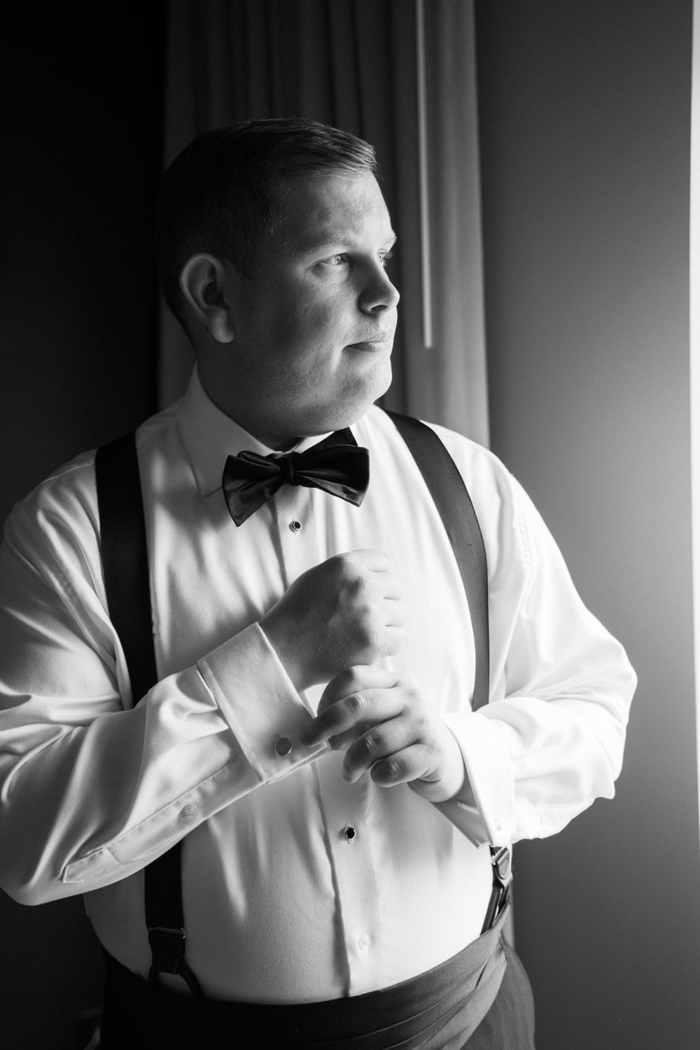 Black and white portrait of a groom in suspenders and bow tie adjusting his cuff link by a window.