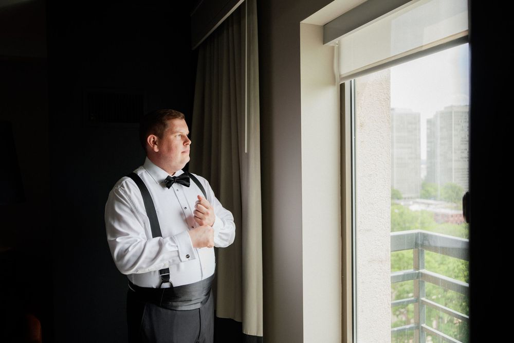 Someone in suspenders and white shirt gazes thoughtfully through window of city apartment.
