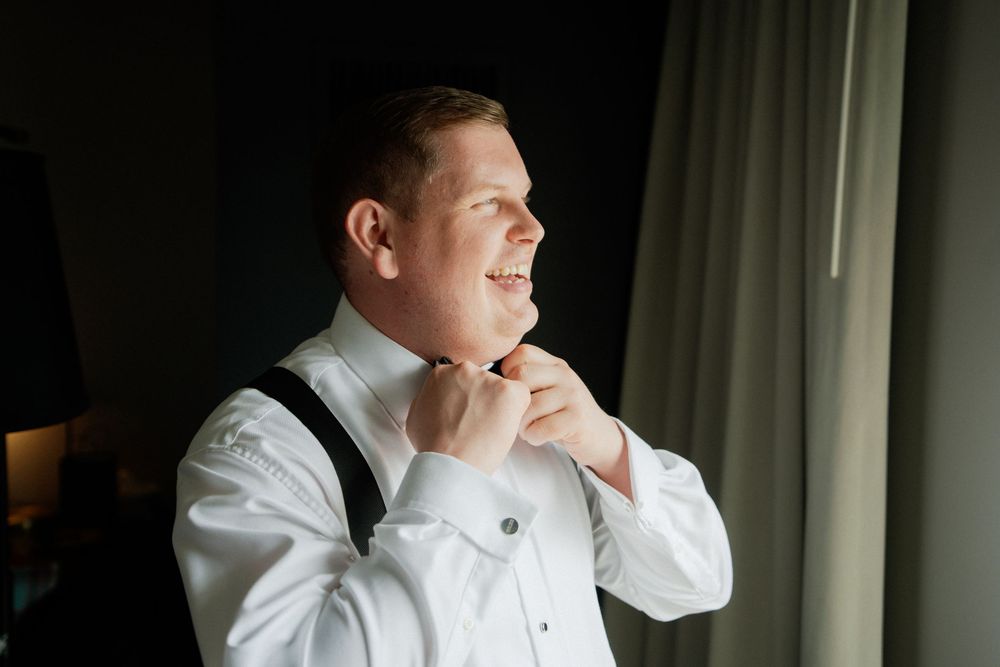 A man in formal attire adjusts his bowtie while getting ready, wearing a crisp white dress shirt with suspenders.