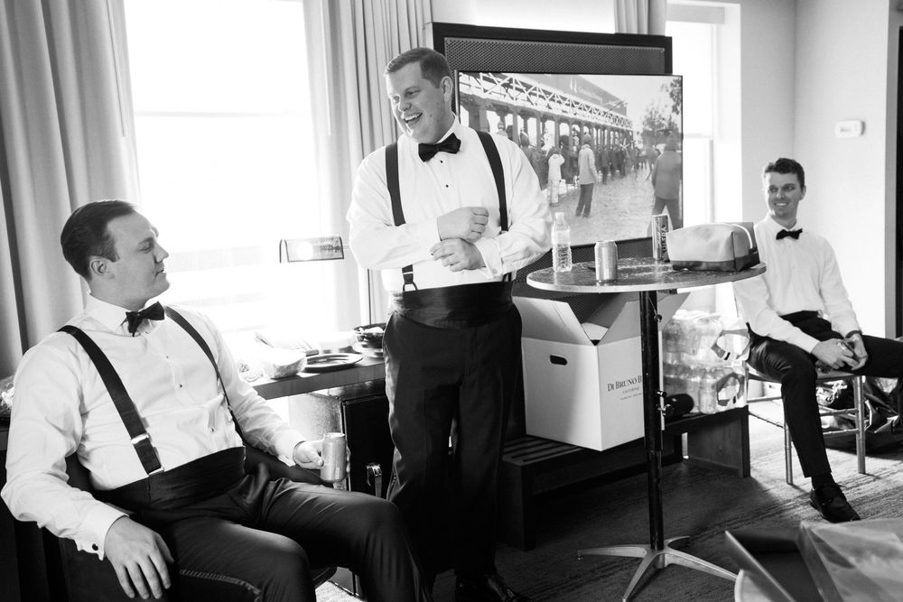 Best men in black bowties and suspenders get ready together before a wedding in a brightly lit room.