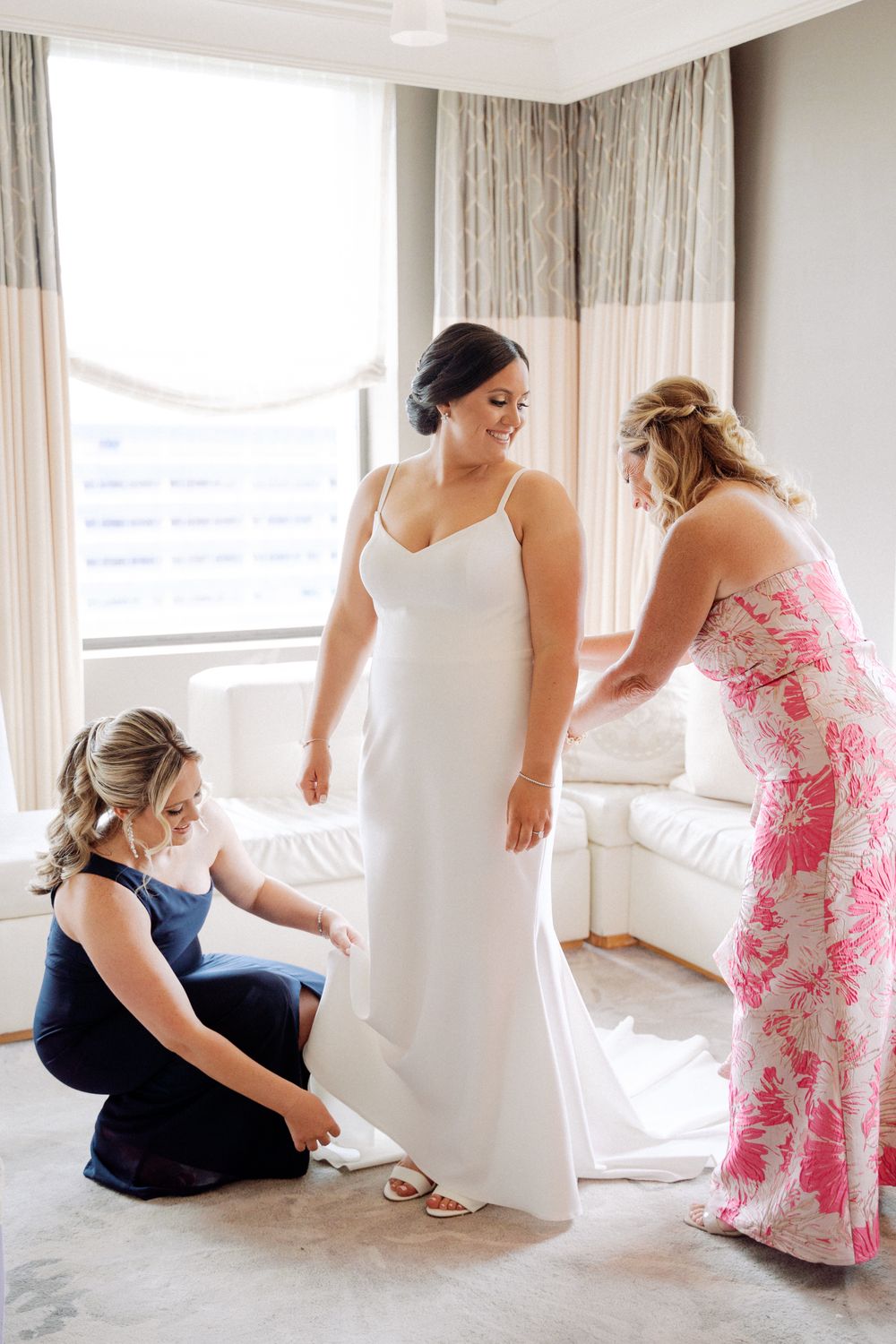 Two women in floral and navy dresses help adjust the hem of a classic white wedding gown in a sunlit room.