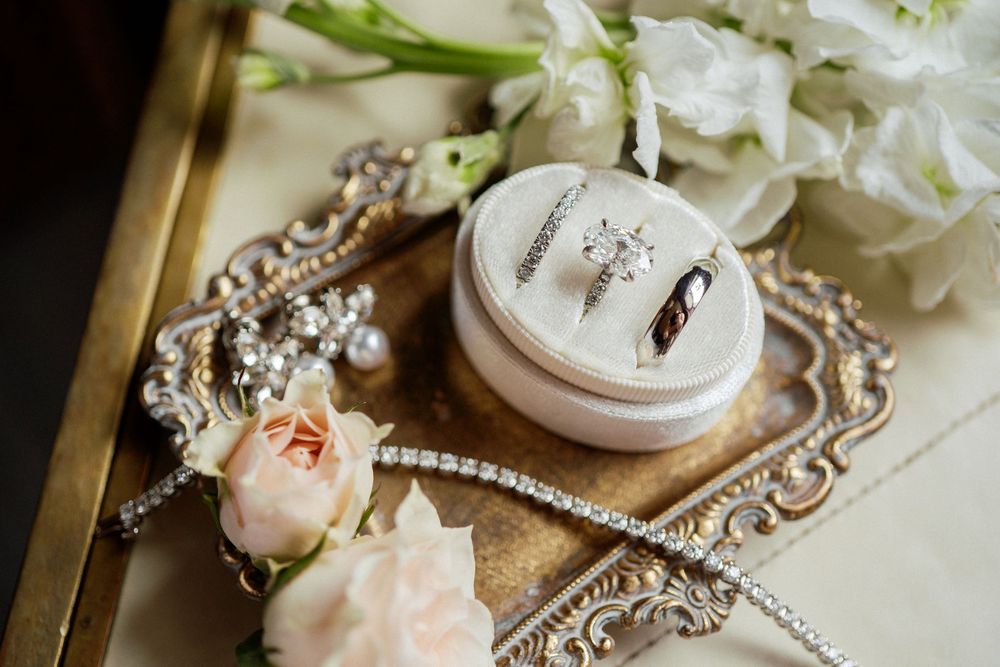 Wedding rings rest in a delicate pink holder on an ornate silver tray with blush roses and white flowers.