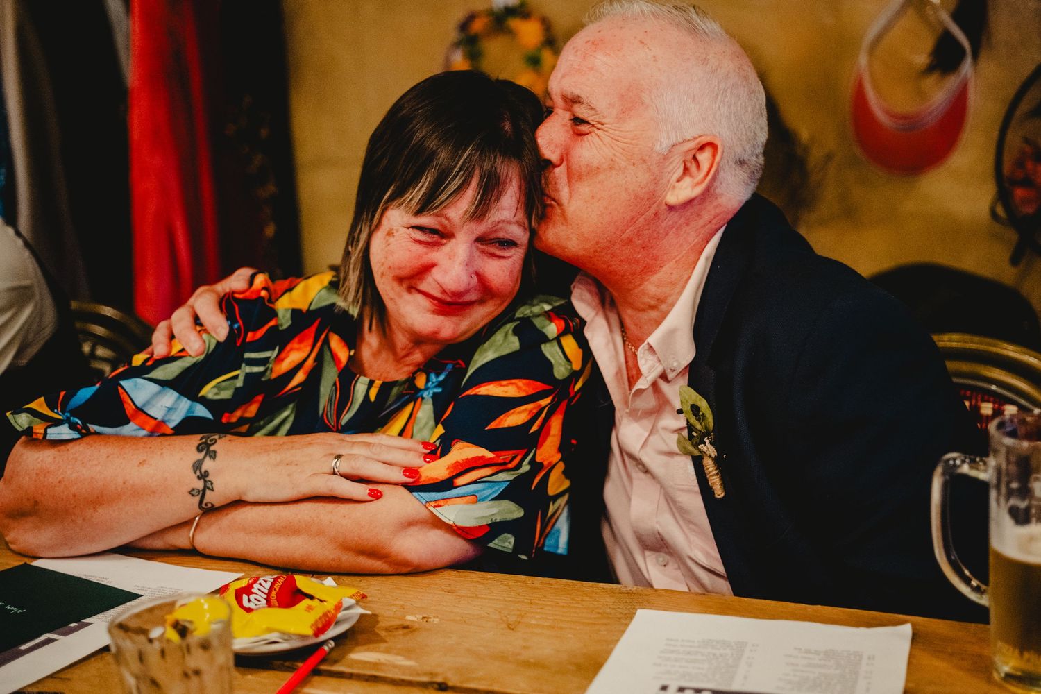 Two people share a tender moment while sitting at a restaurant table with plates of food and drinks.