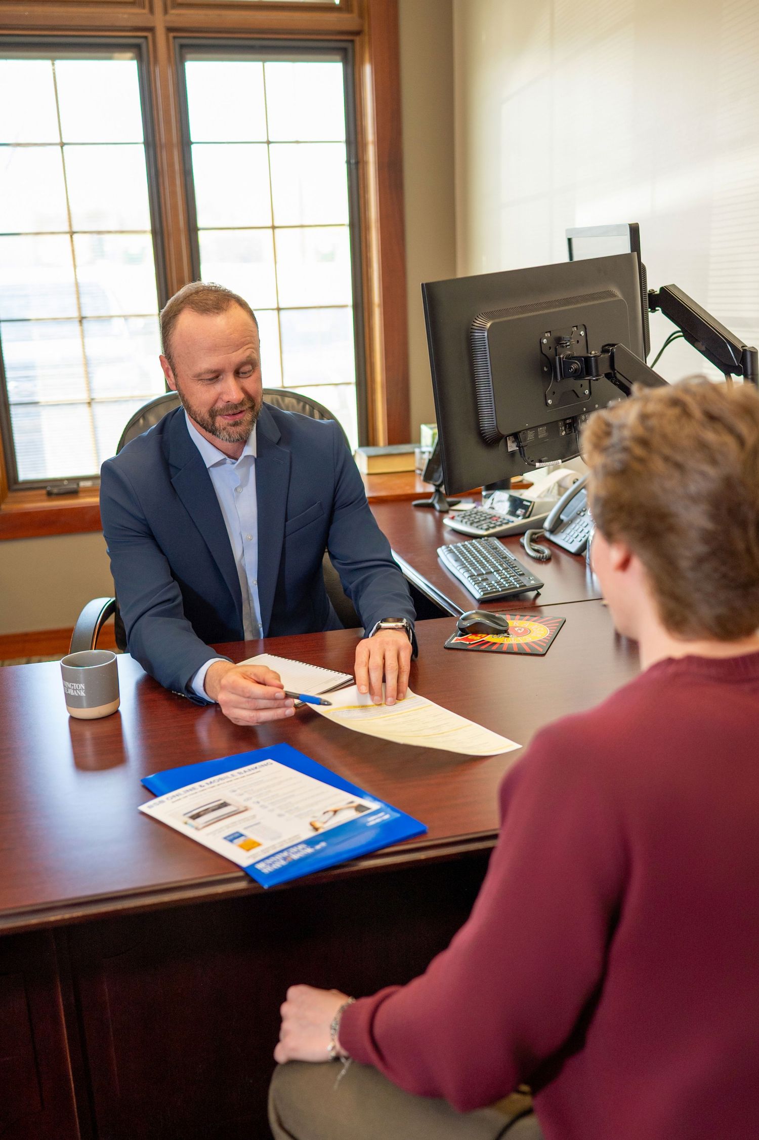 Two people meet in an office setting at a desk with papers and a computer monitor visible in the background.