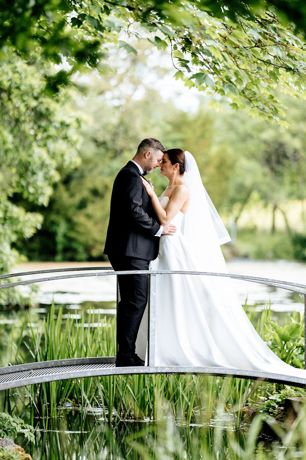 Newlywed couple stands intimately on arched garden bridge surrounded by lush greenery and sunlight.