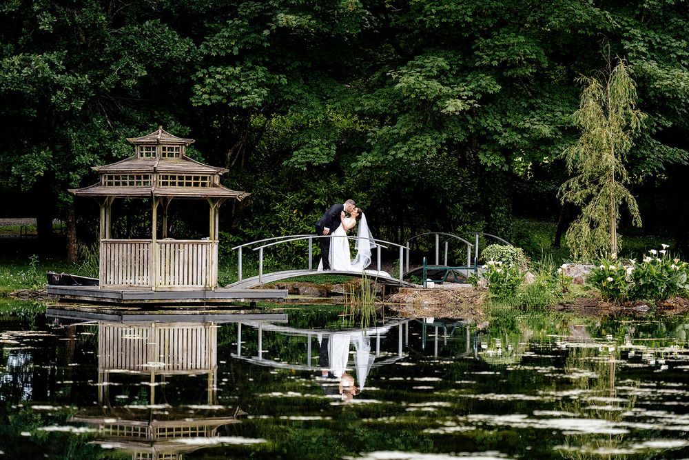 A couple stands on a bridge near a gazebo at a serene pond with perfect reflections in a garden setting.