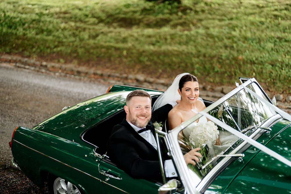 A couple rides together in a vintage green classic car decorated with wedding ribbons and bows on a sunny day.
