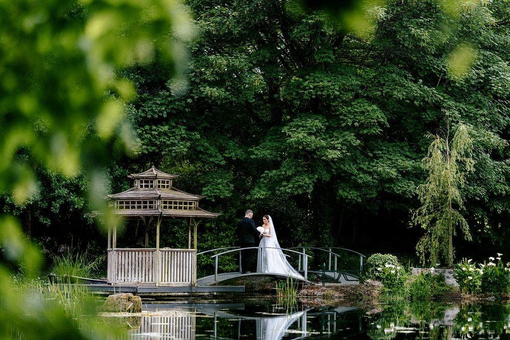 A romantic wedding scene at a serene garden gazebo overlooking a reflective pond surrounded by lush green trees.