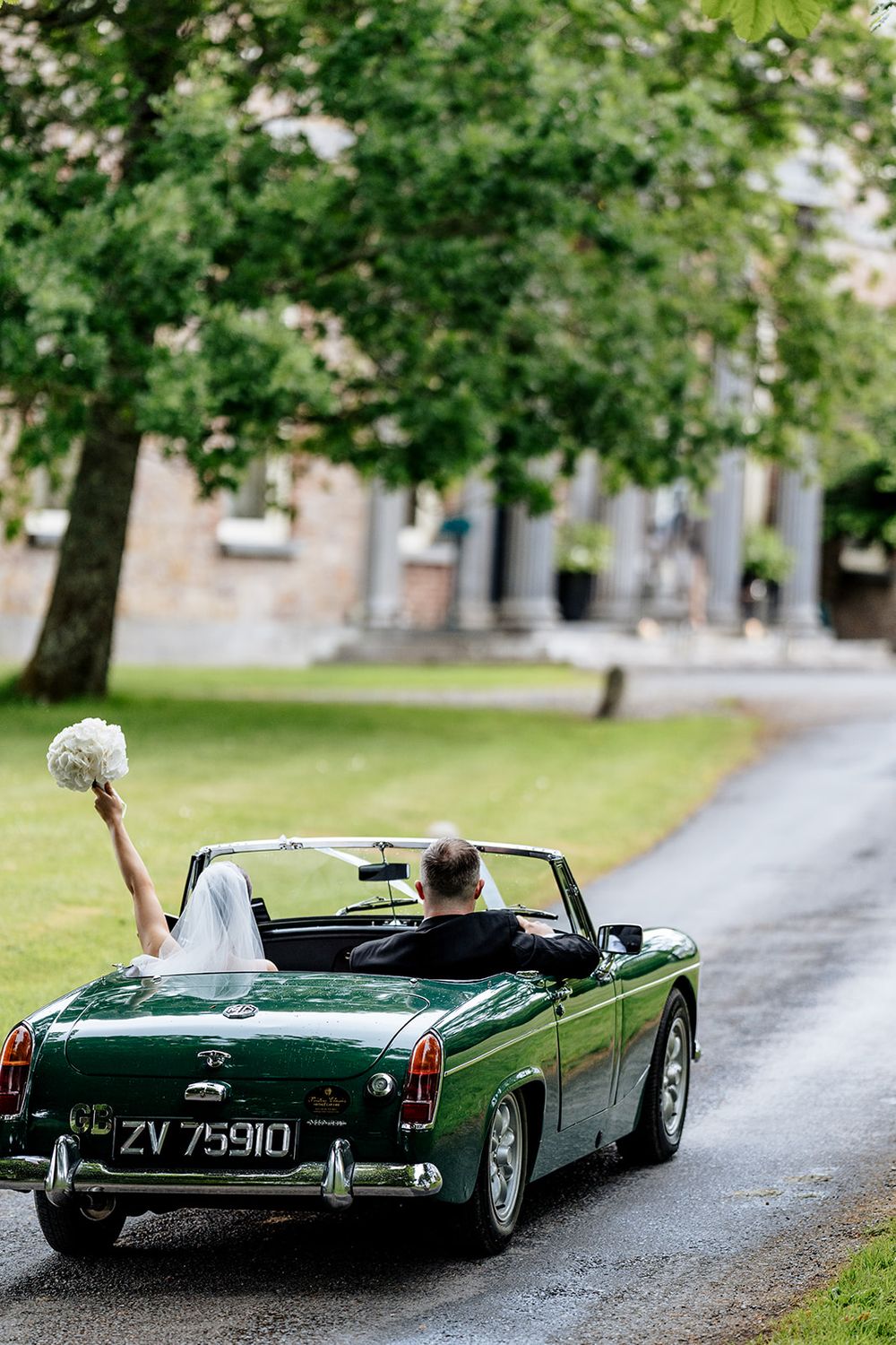A green vintage MG convertible drives down a tree-lined path as a bouquet is held up from the passenger seat.