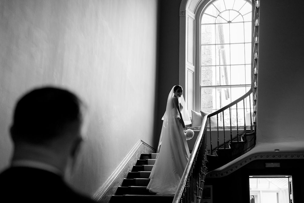 A black and white wedding photo capturing a bride ascending a grand staircase near an arched window, silhouetted figure watching.
