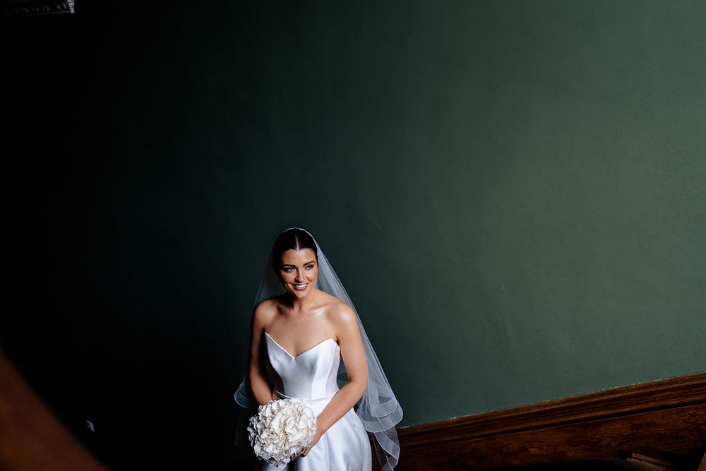 A bride in a white wedding gown holding a bouquet sits elegantly against a dark green wall.
