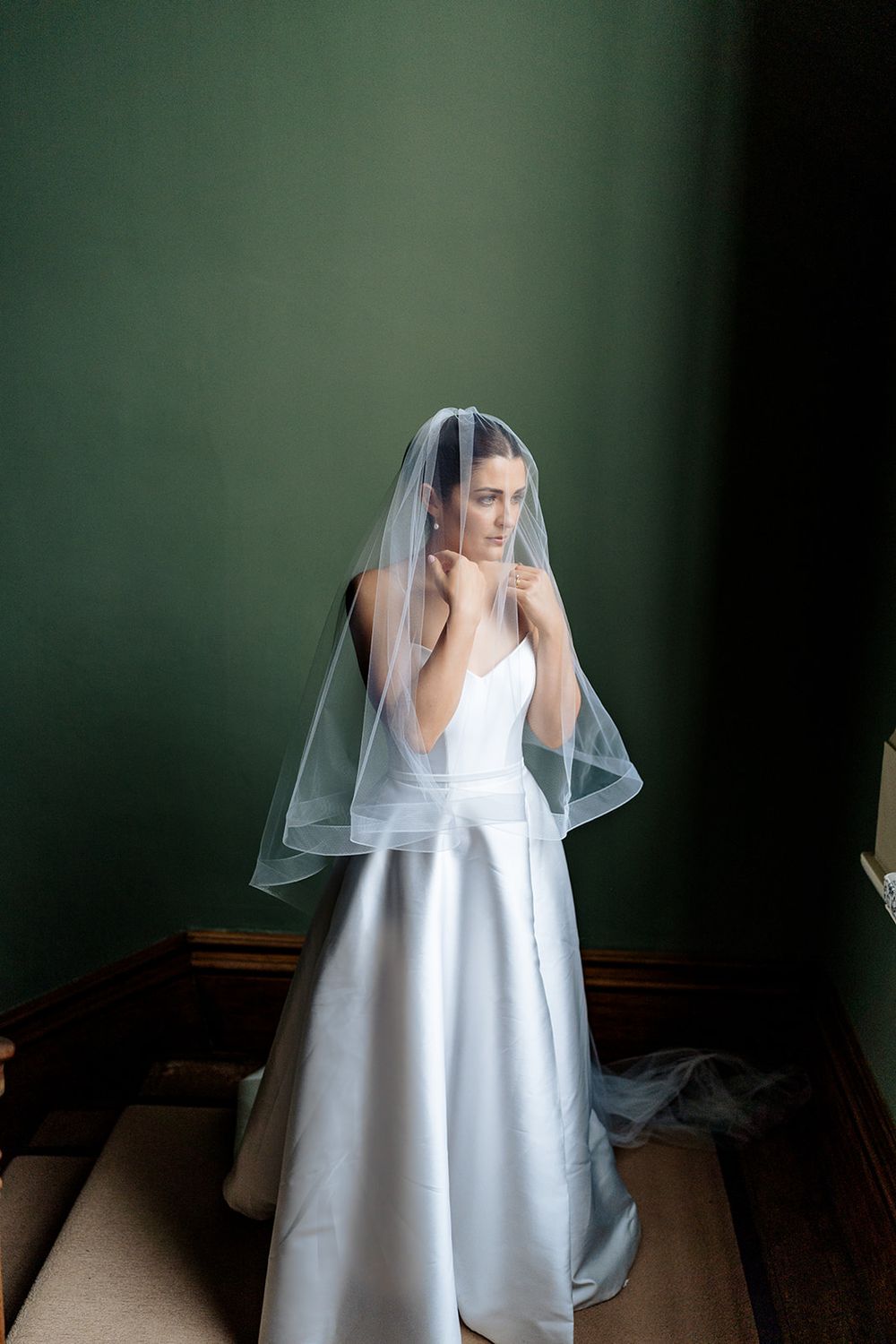 A bride in an elegant white satin wedding gown and sheer veil stands thoughtfully against a dark green wall background.