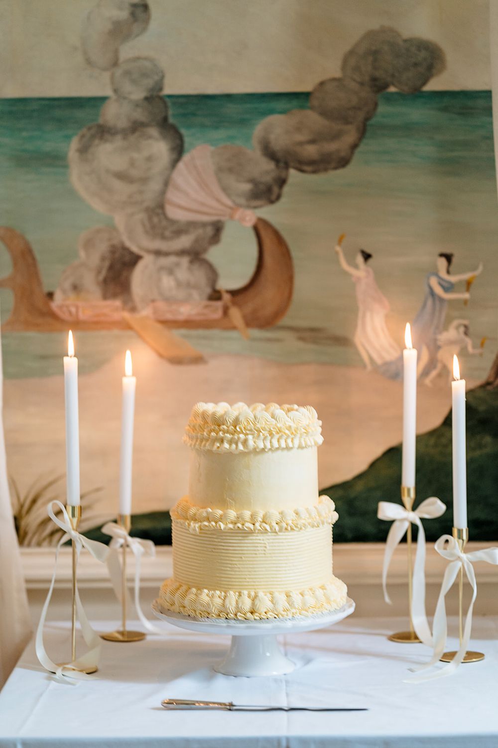 Elegant two-tier cream wedding cake with delicate piping sits on white tablecloth with tall taper candles and painted wall backdrop.