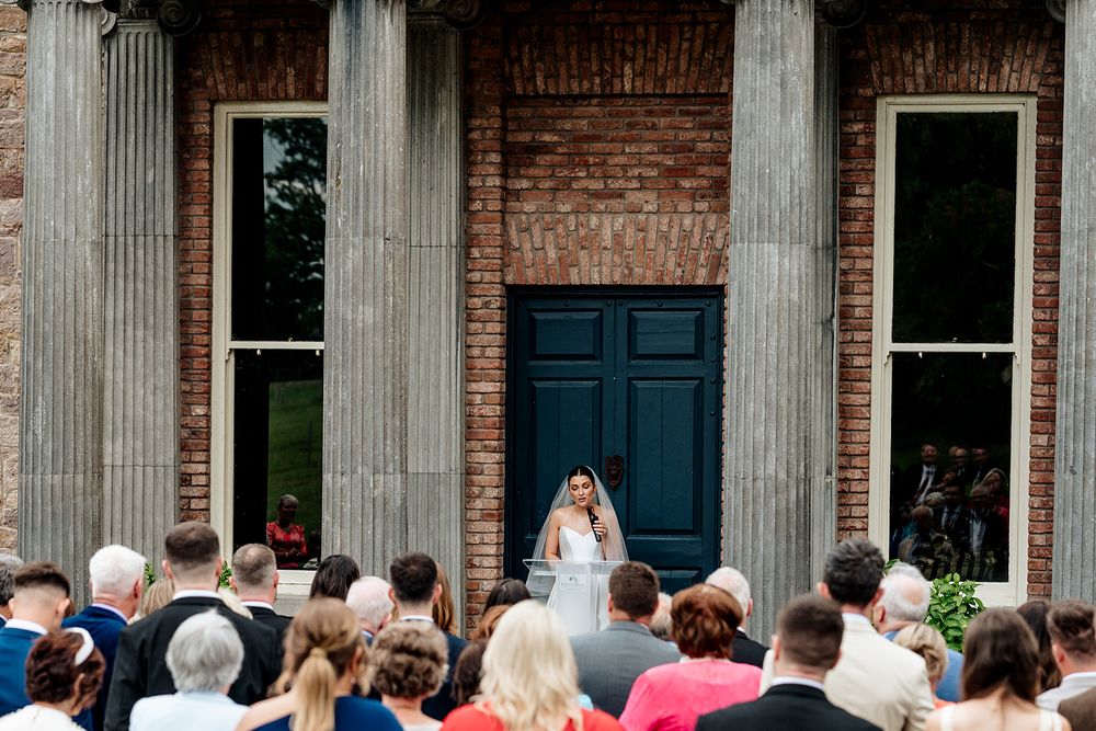 A crowd gathers in front of a historic brick building with columns to watch a wedding ceremony at the entrance.