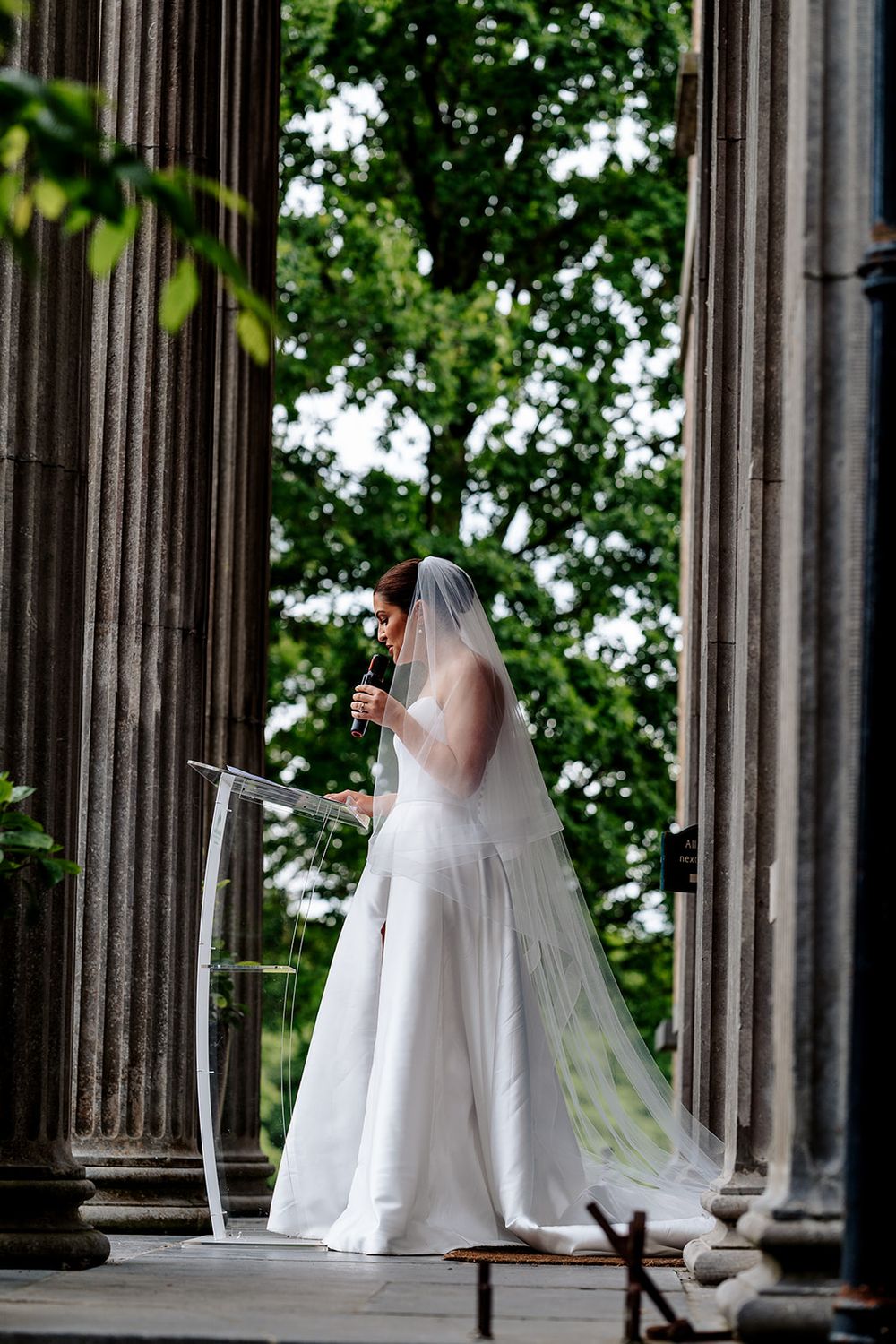 A bride in a flowing white wedding gown and veil stands between classical stone columns framed by green foliage.