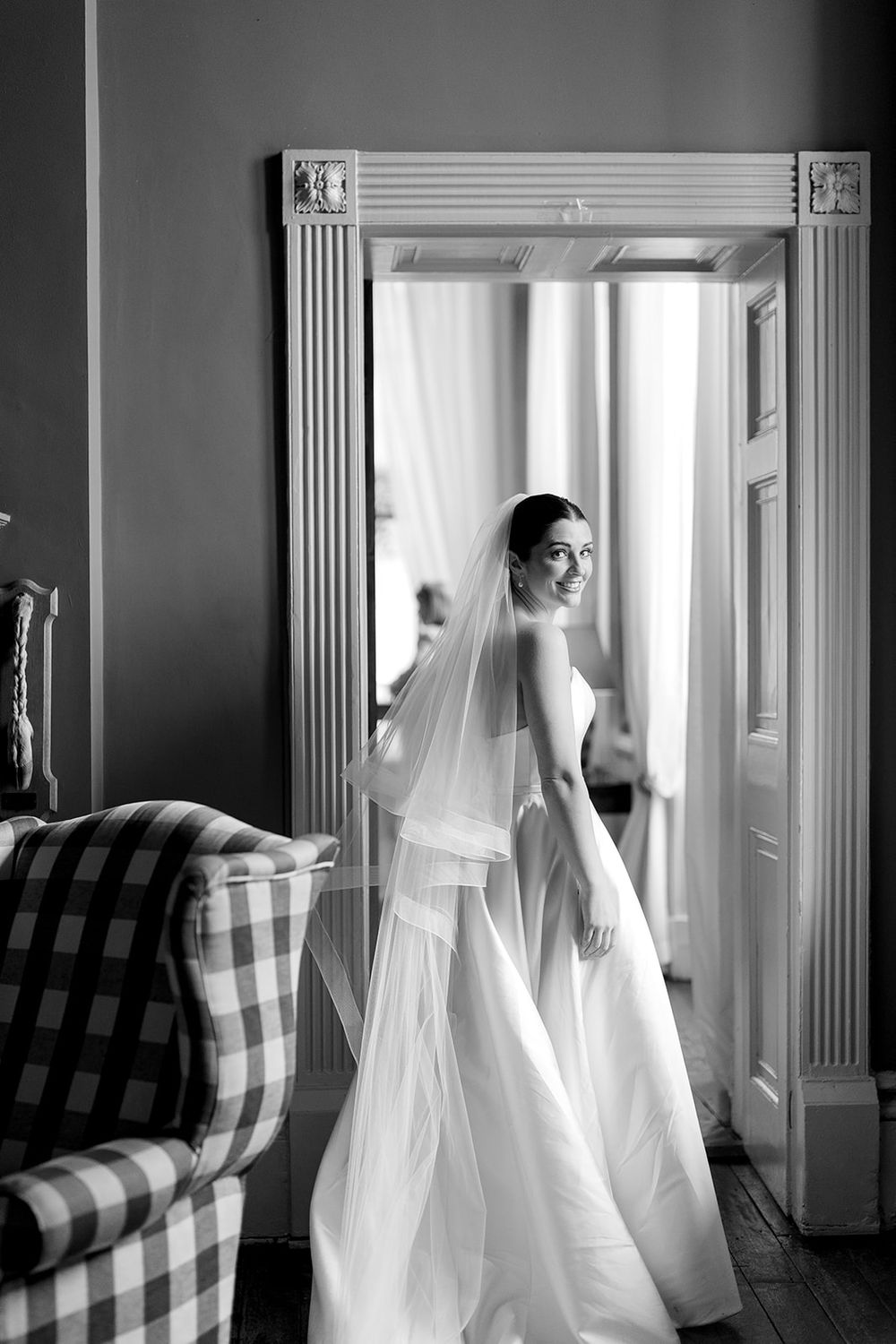 A bride in an elegant white wedding gown and veil poses beside an ornate doorframe in black and white.