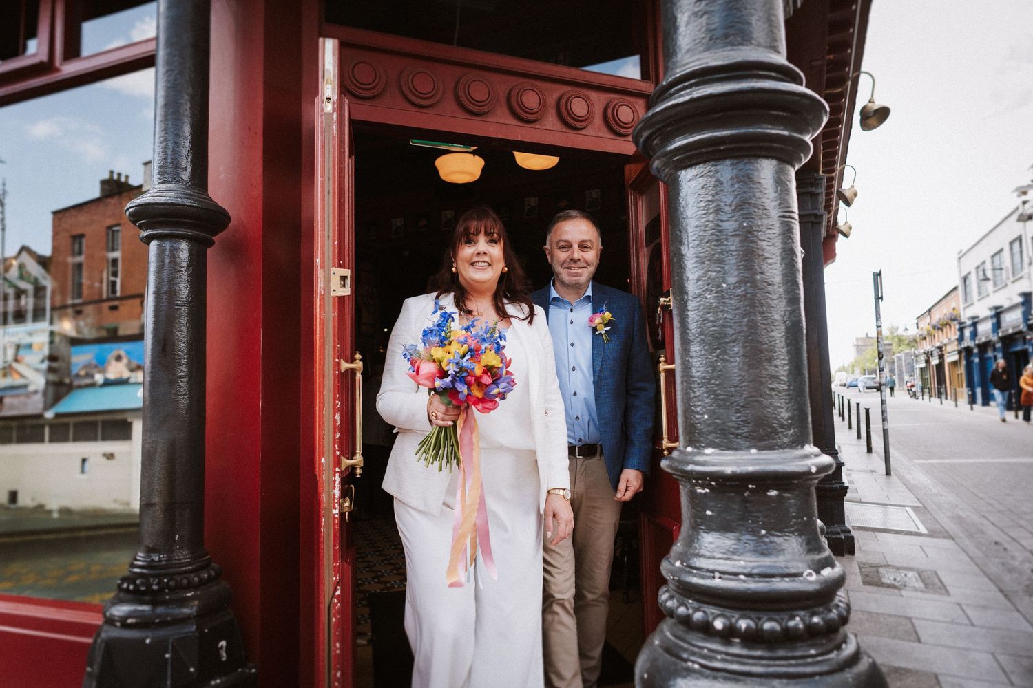 Happy couple exits red doorway of historic building with ornate cast iron posts on city street.