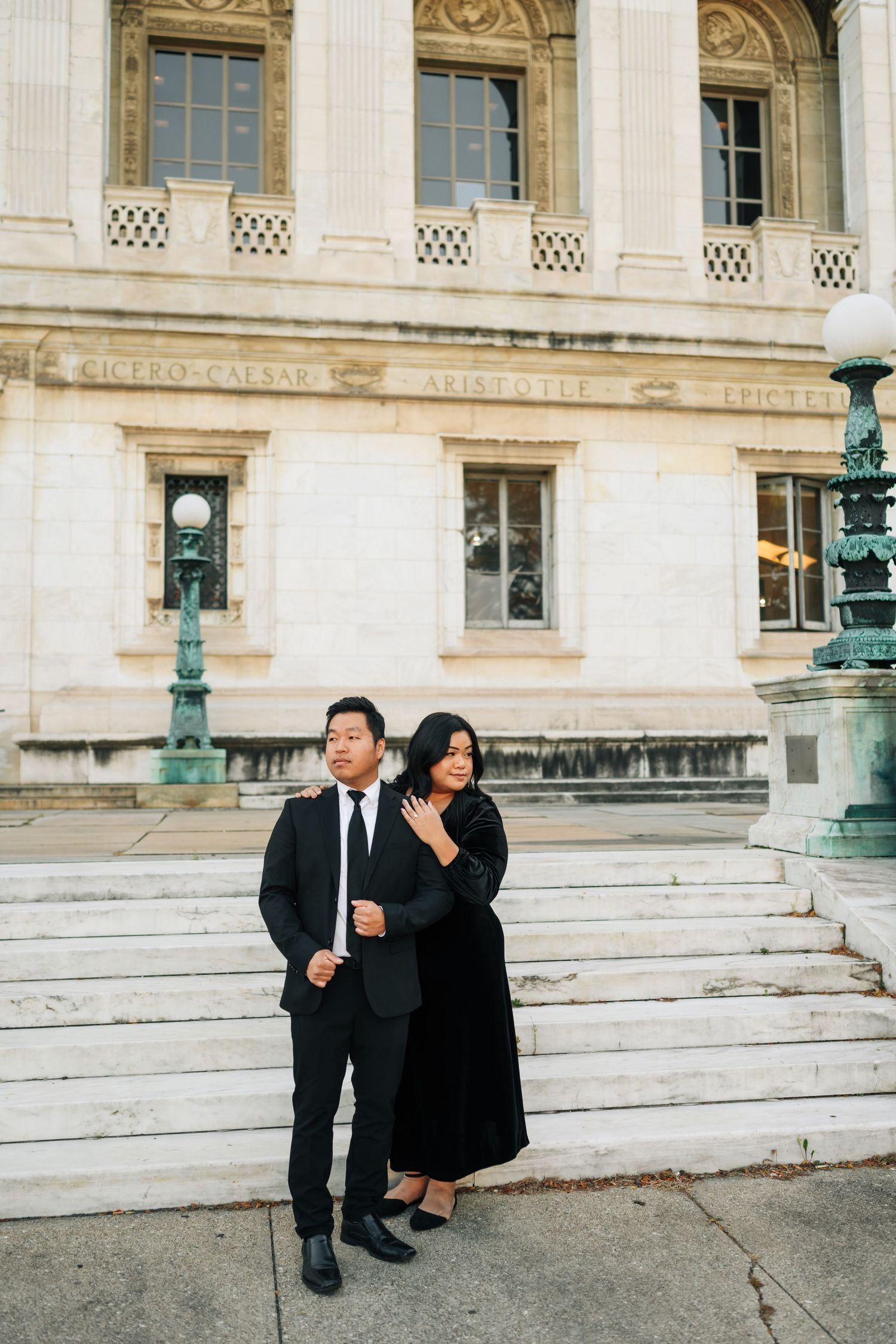 Elegant couple in formal black attire posing on stone steps of a grand historic building with ornate architecture.