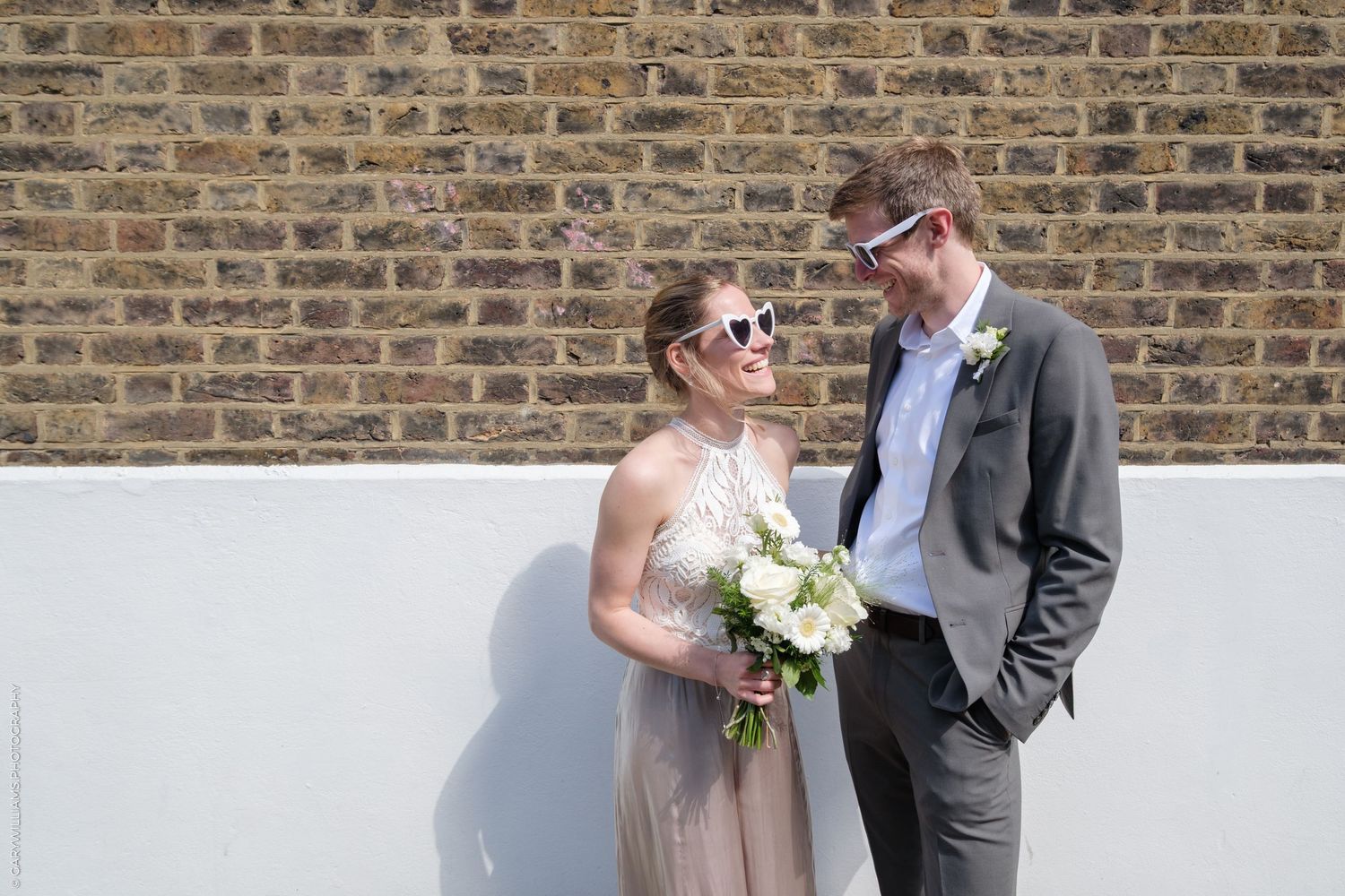 A couple in formal attire poses in a romantic narrow alleyway with brick walls and greenery in the background.