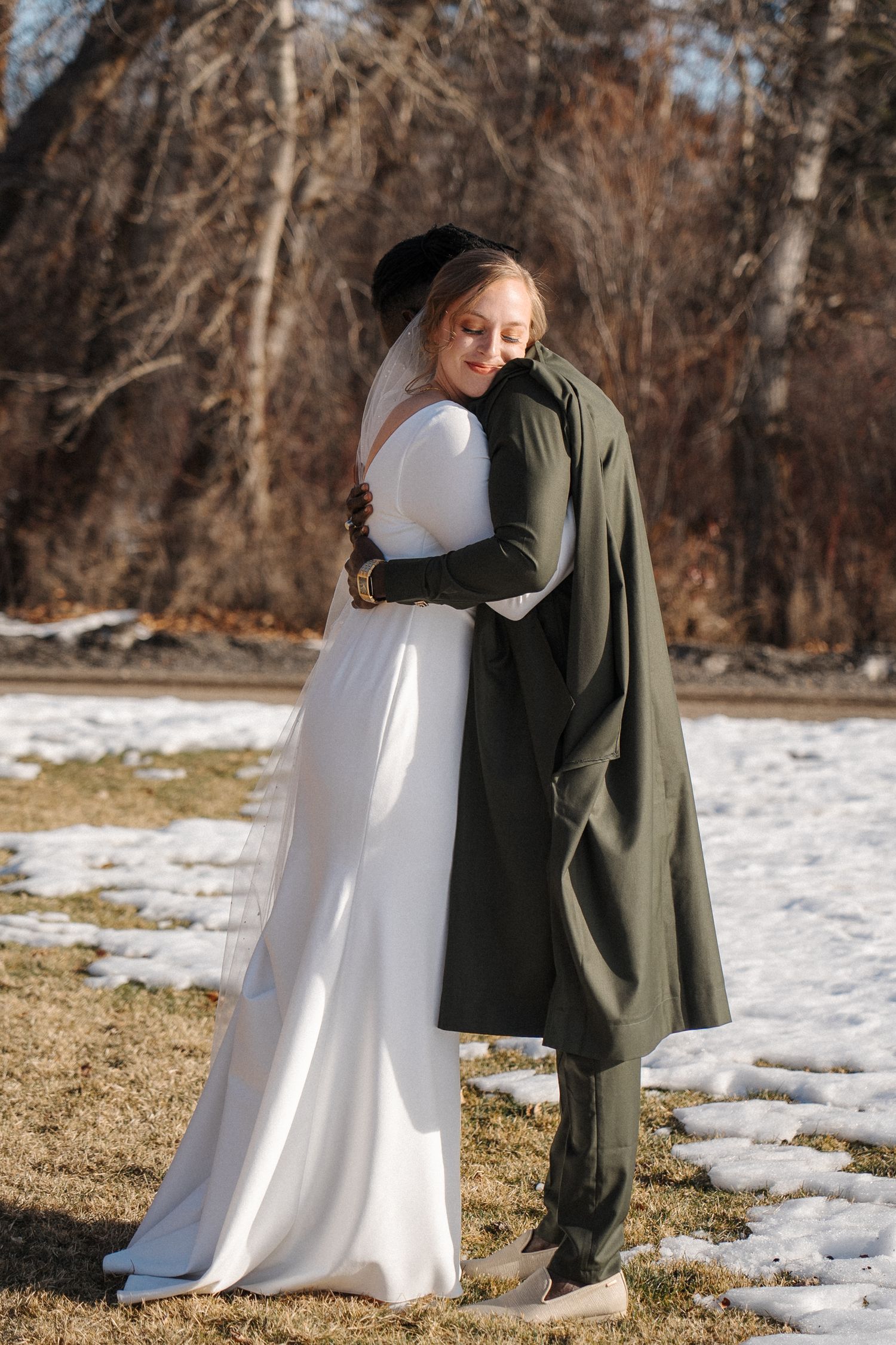 A couple embraces outdoors on a snowy winter day, with the bride in white and partner wearing a dark green coat.